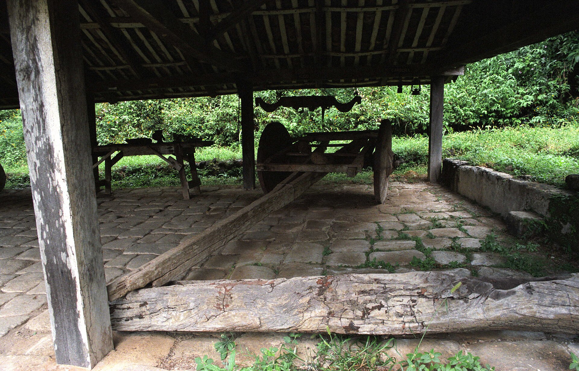 An open wooden shed with a tiled roof and stone floor sheltering an old wooden cart and a long timber beam, with green vegetation behind