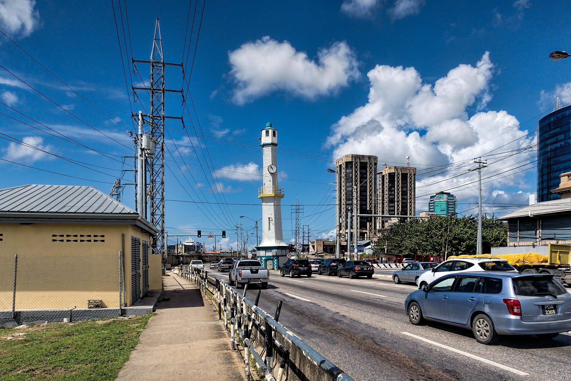 Cars on a multi-lane road pass a white and yellow lighthouse tower with a clock, with high-rise towers and power lines under a blue sky in Port of Spain