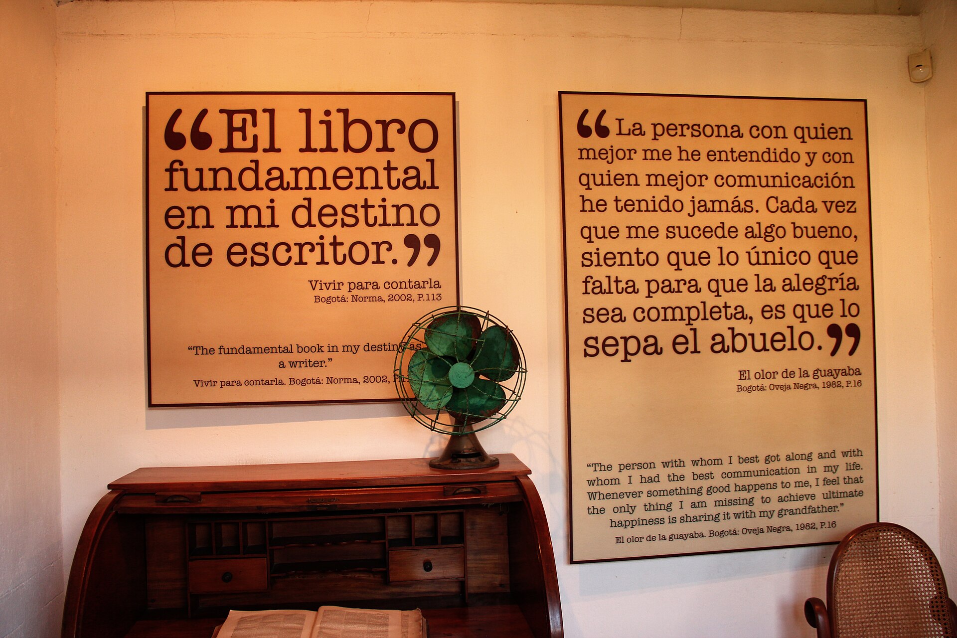 A room with an old roll-top desk, a green electric fan and two large wall panels quoting García Márquez in Spanish and English