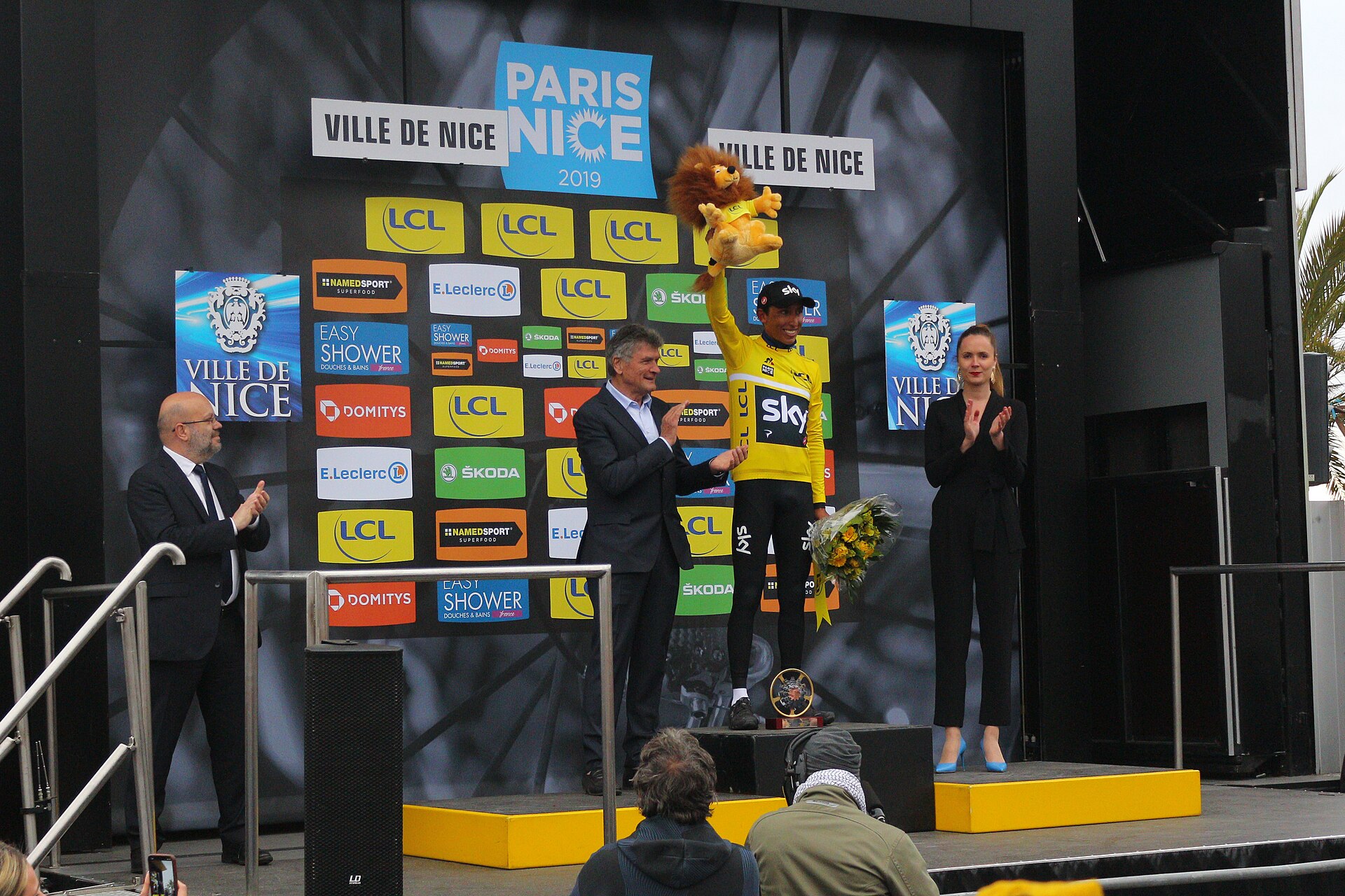 Egan Bernal in a yellow leader's jersey raising a lion mascot on a podium in Nice, with officials applauding