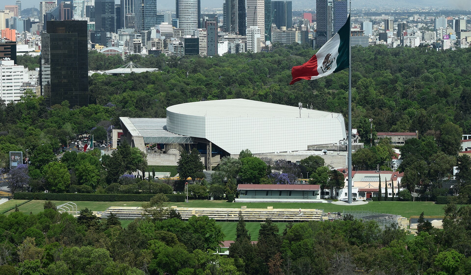 A giant Mexican flag on a tall pole above green parkland and a white curved-roof building, with Mexico City towers on the skyline