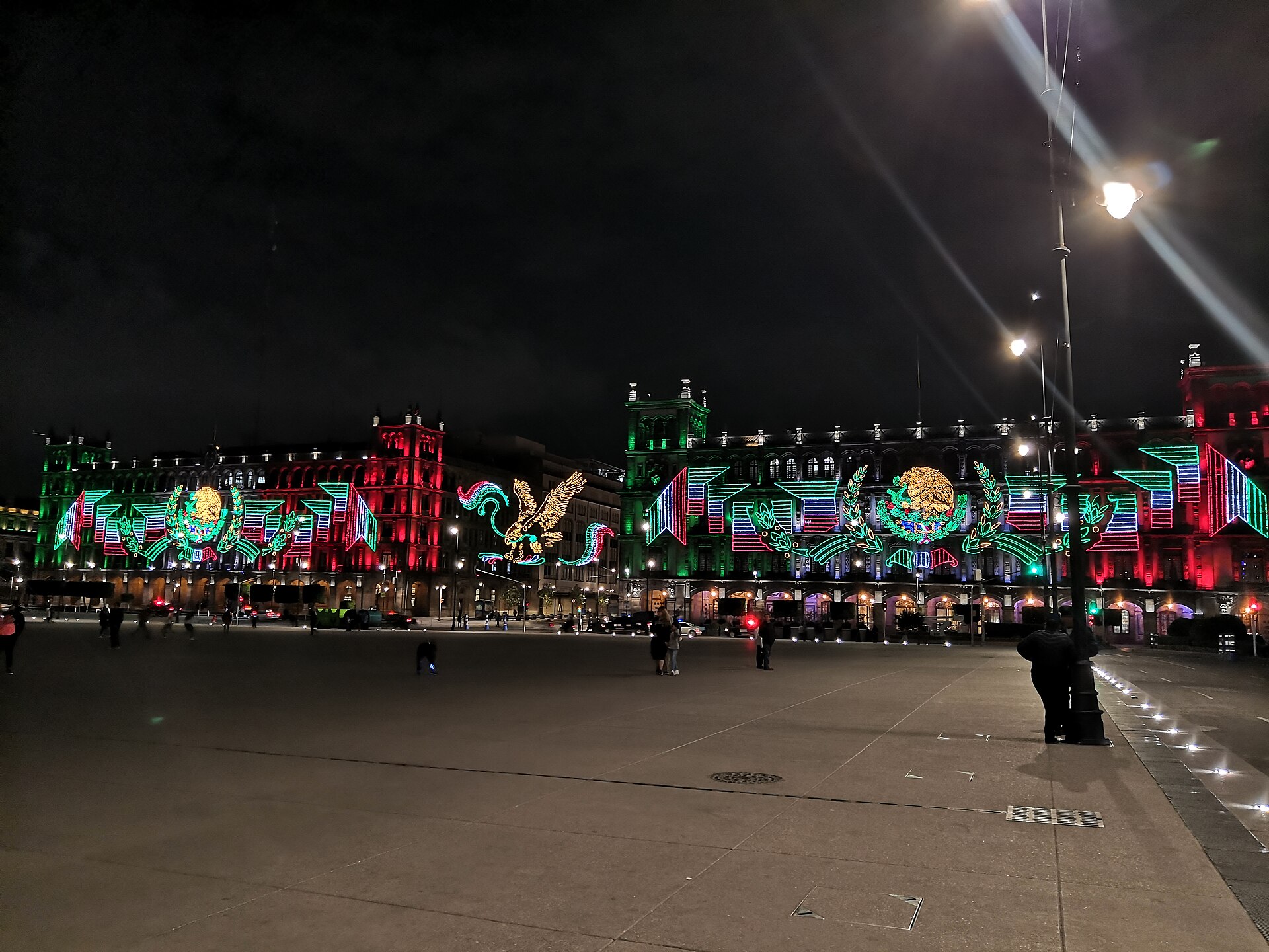 Night view across the Zócalo to buildings lit with green, white and red patriotic decorations including eagle designs