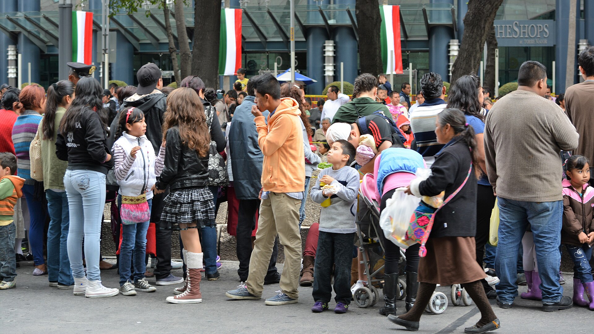 Families and children stand along a pavement on Paseo de la Reforma beneath Mexican flags