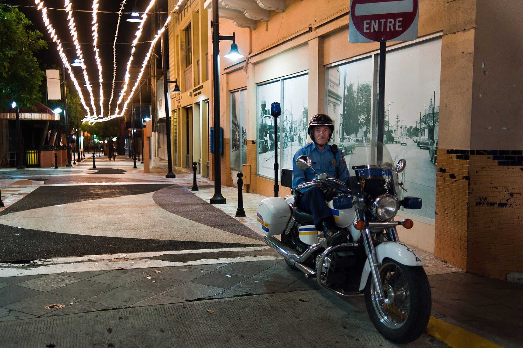 A Puerto Rico Police officer in a helmet and blue uniform sits on a police motorcycle on a lit pedestrian street at night in Mayagüez