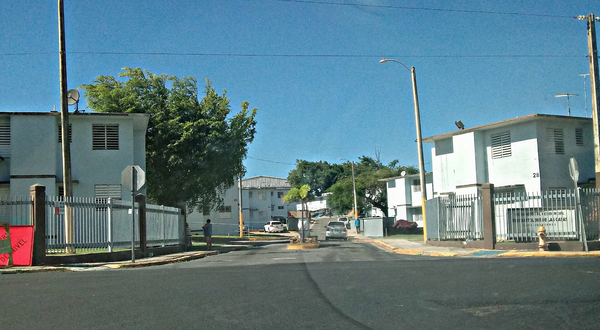 A street between low white public housing blocks with fences, trees and parked cars under a blue sky at Residencial Las Casas in Santurce, San Juan
