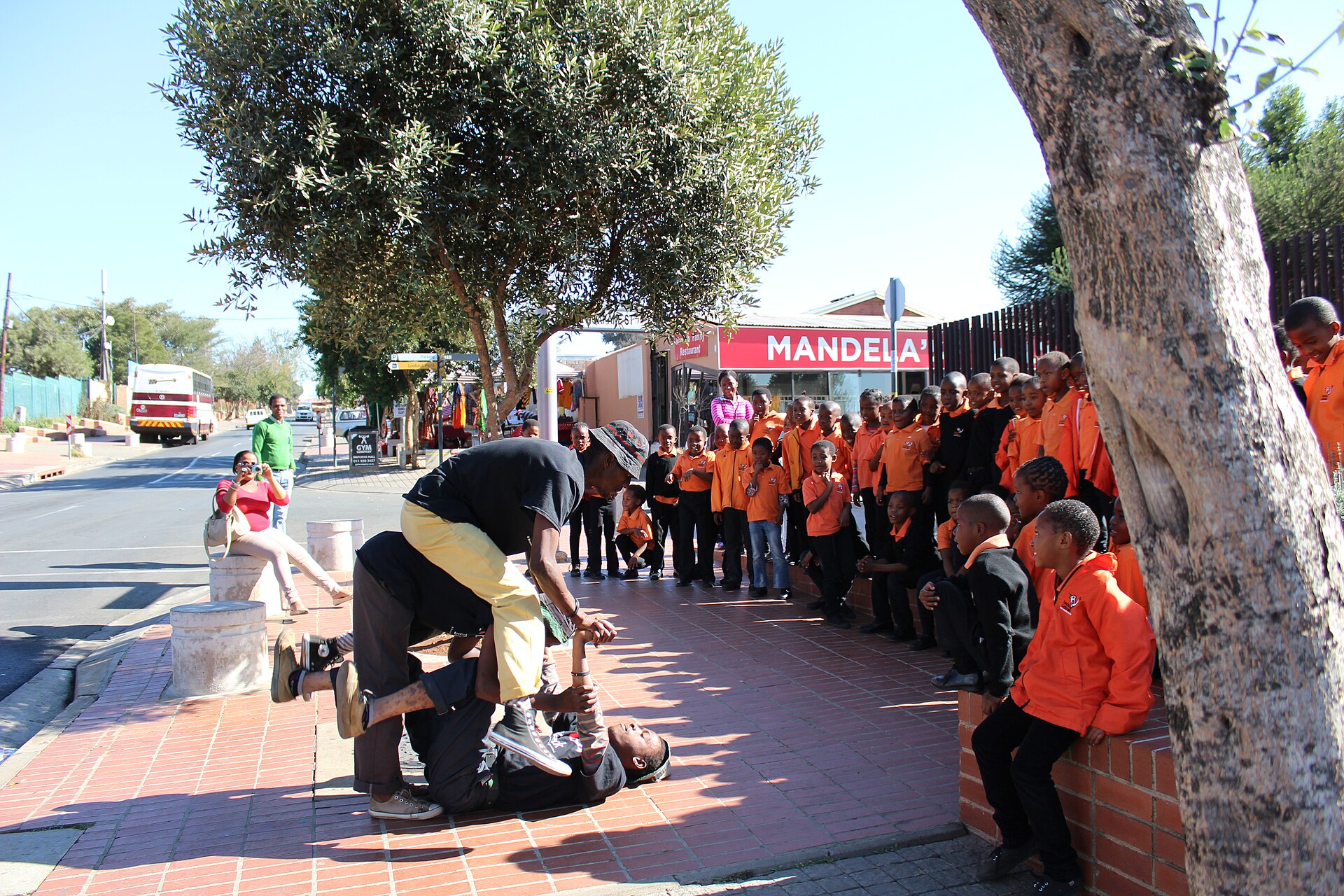 Two street performers do acrobatics on a brick pavement while schoolchildren in orange jackets watch, with a shop sign reading Mandela behind them