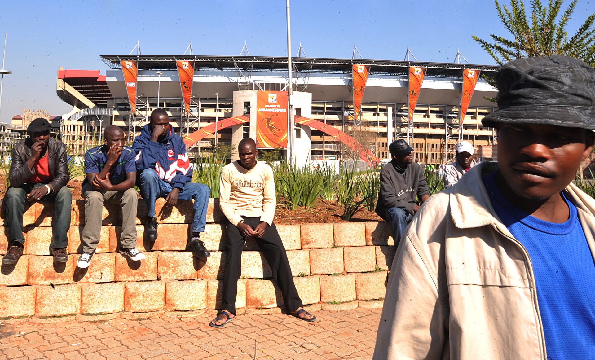 Men sit on a low brick wall in front of Ellis Park Stadium, decorated with orange World Cup banners, in Johannesburg