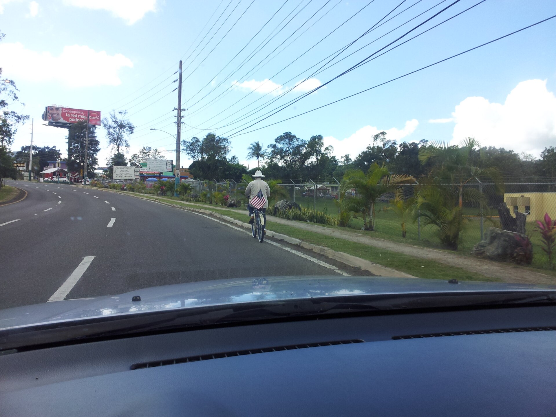 A cyclist in a hat rides beside a road in Cayey, Puerto Rico, seen through a car windscreen, with palm trees and a billboard behind