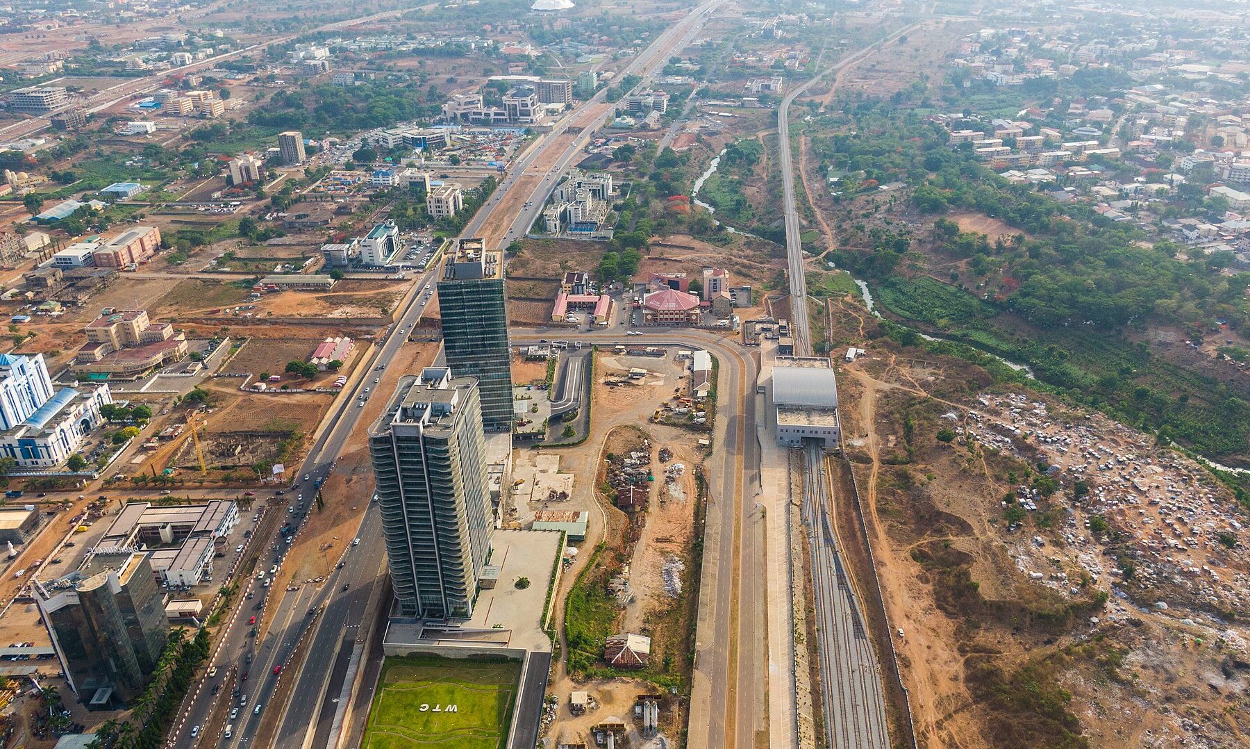 Aerial view of the Central Business District of Abuja, with two tall glass towers beside a lawn marked WTC, wide highways, a rail line and low-rise districts stretching to the horizon