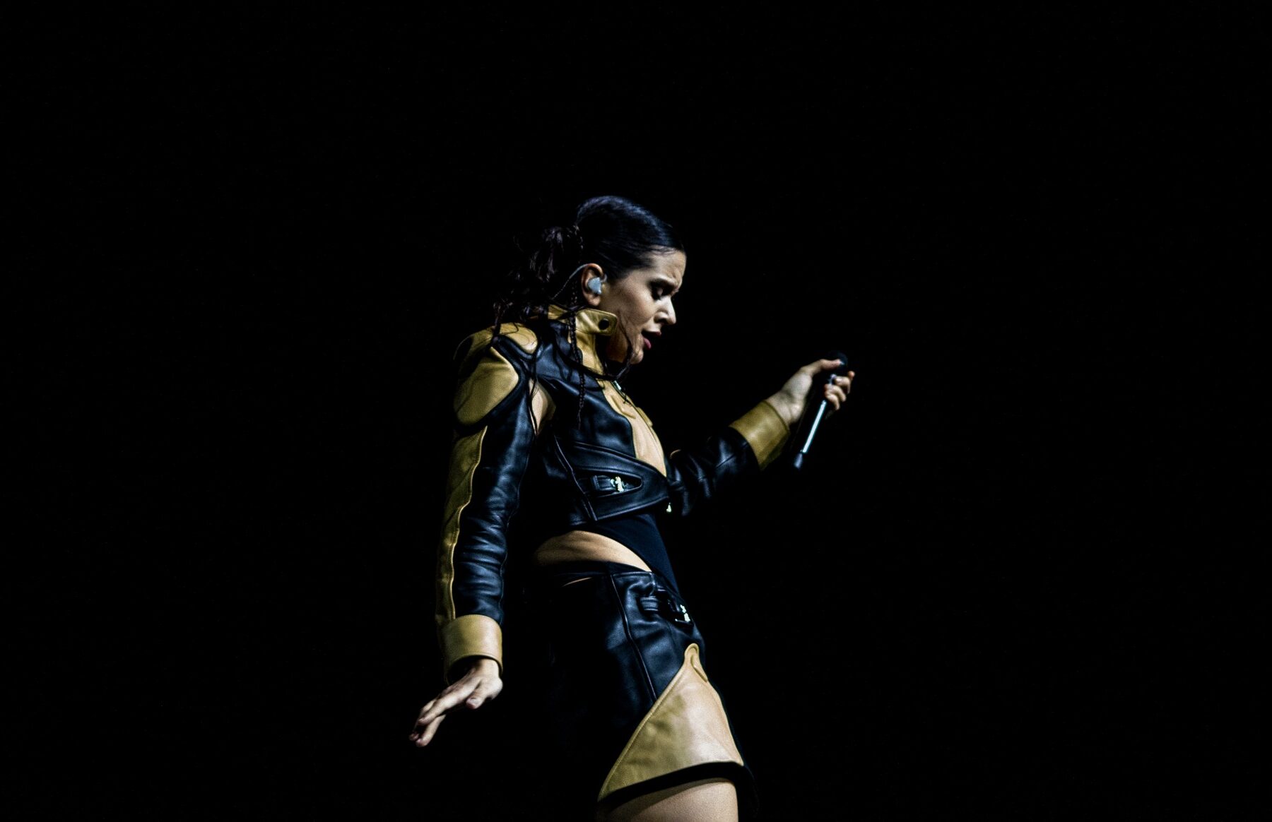 Rosalía singing on a dark stage in a black and gold leather outfit, microphone in hand