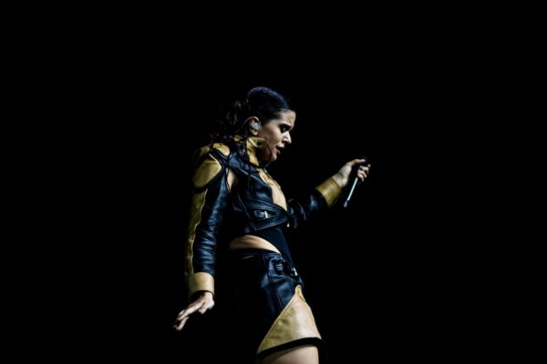 Rosalía singing on a dark stage in a black and gold leather outfit, microphone in hand