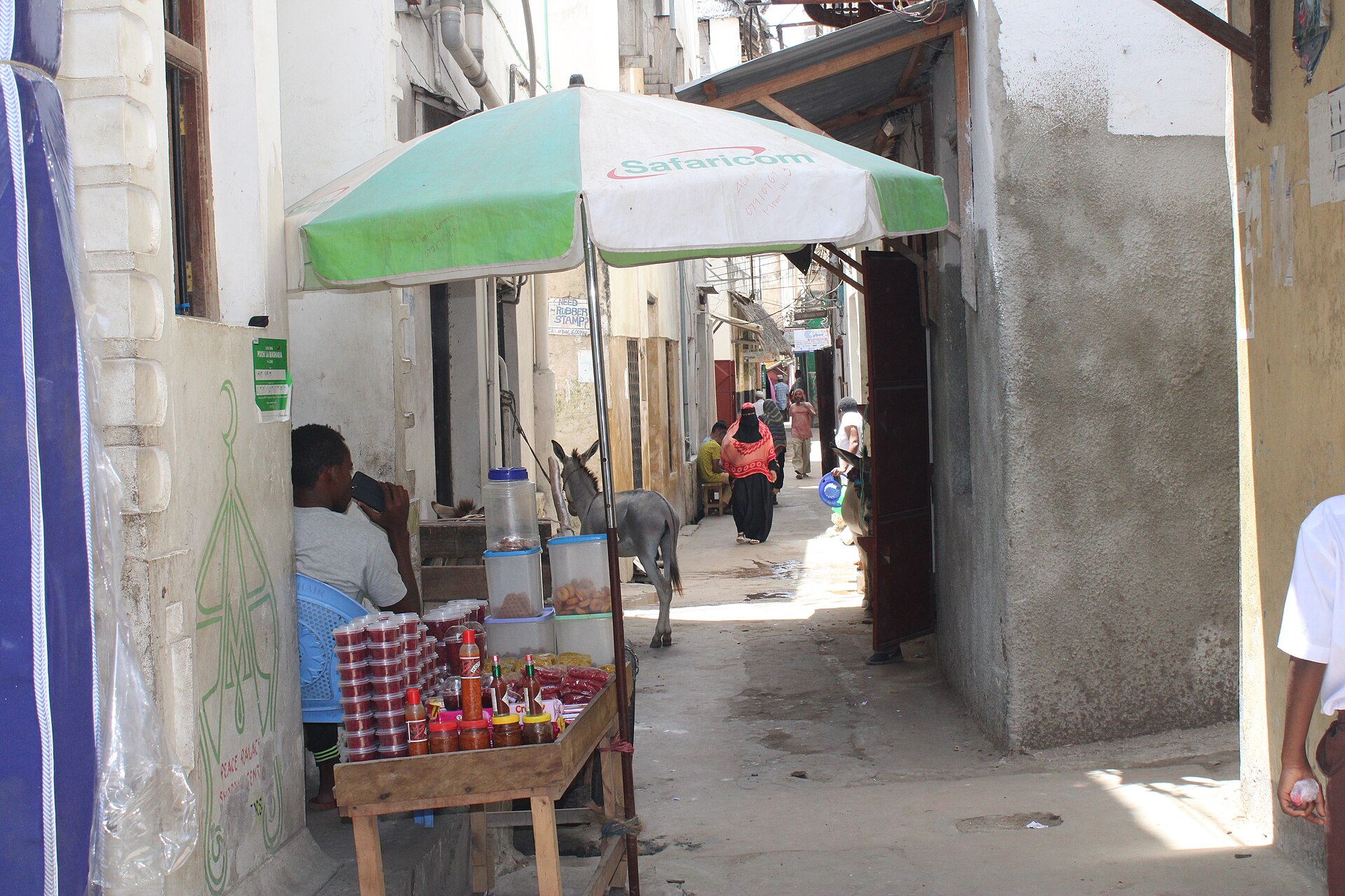A narrow whitewashed alley in Lamu Old Town with a street stall under a green umbrella, a donkey and a woman in a headscarf walking away