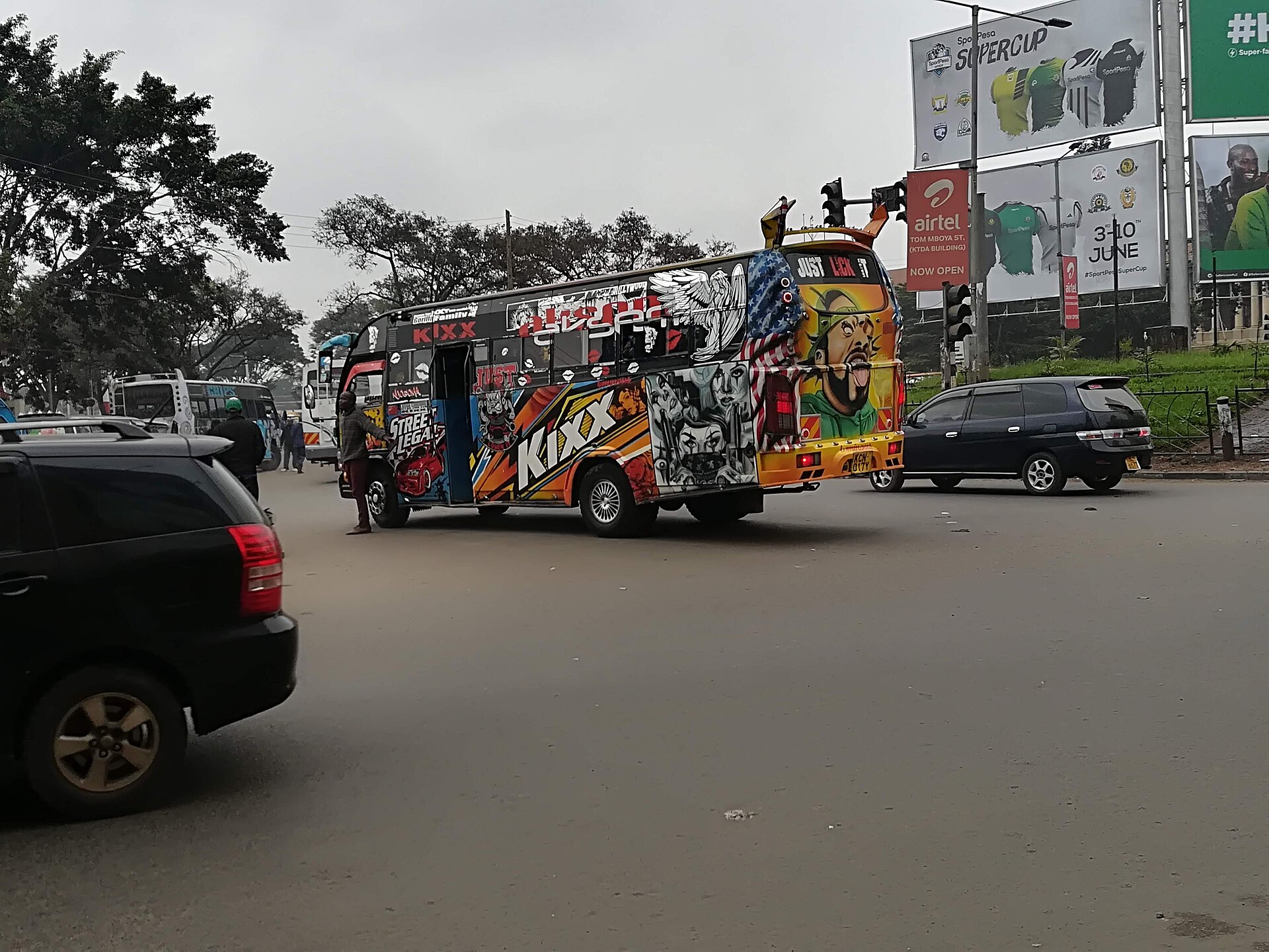 A matatu minibus covered in colourful airbrushed portraits and the word KiXX crosses a busy Nairobi road junction beside billboards