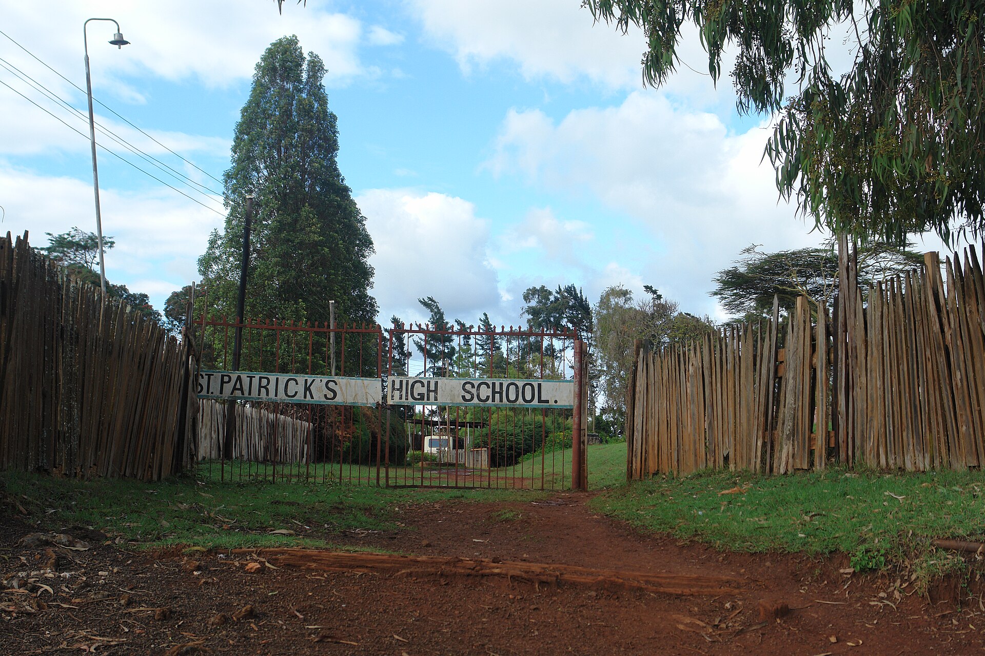 The iron gate of St Patrick's High School in Iten between wooden stake fences, with a red dirt road and tall trees