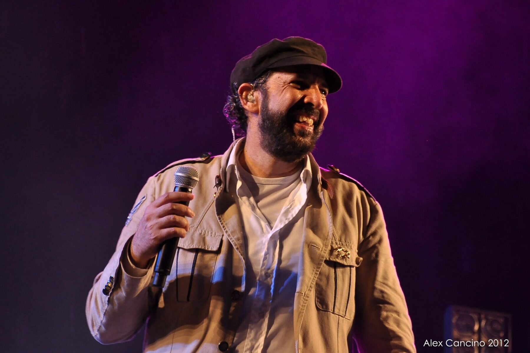 Juan Luis Guerra smiling on stage in a cap and khaki jacket, holding a microphone against a purple-lit background