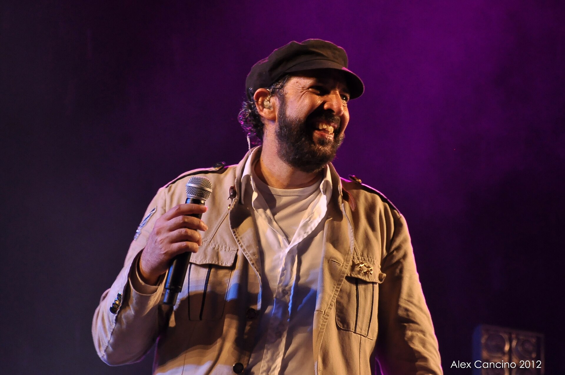 Juan Luis Guerra smiling on stage in a cap and khaki jacket, holding a microphone against a purple-lit background