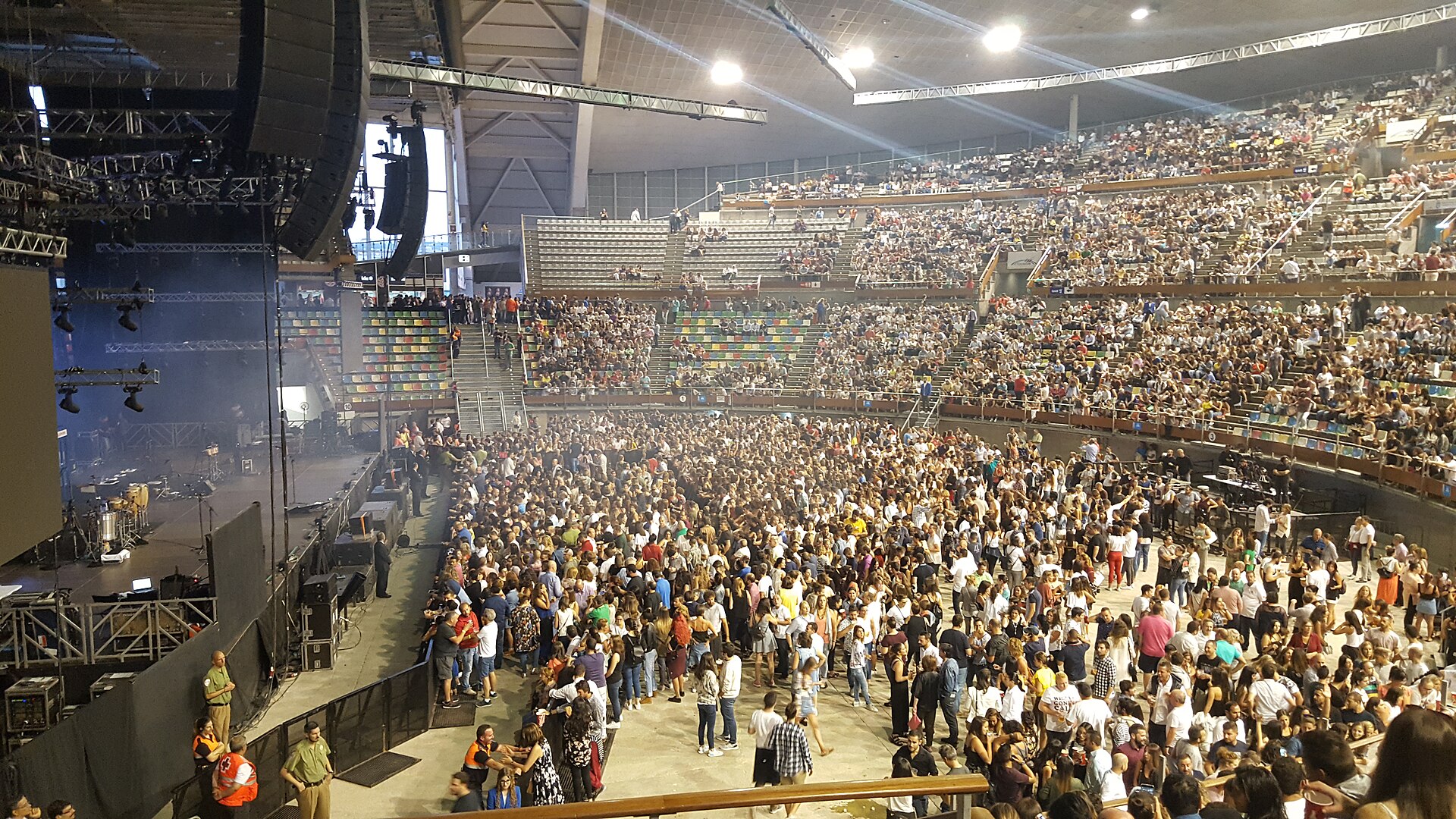 Crowd on the floor and in the stands of an indoor arena before a concert, with stage speakers and lighting rigs at left