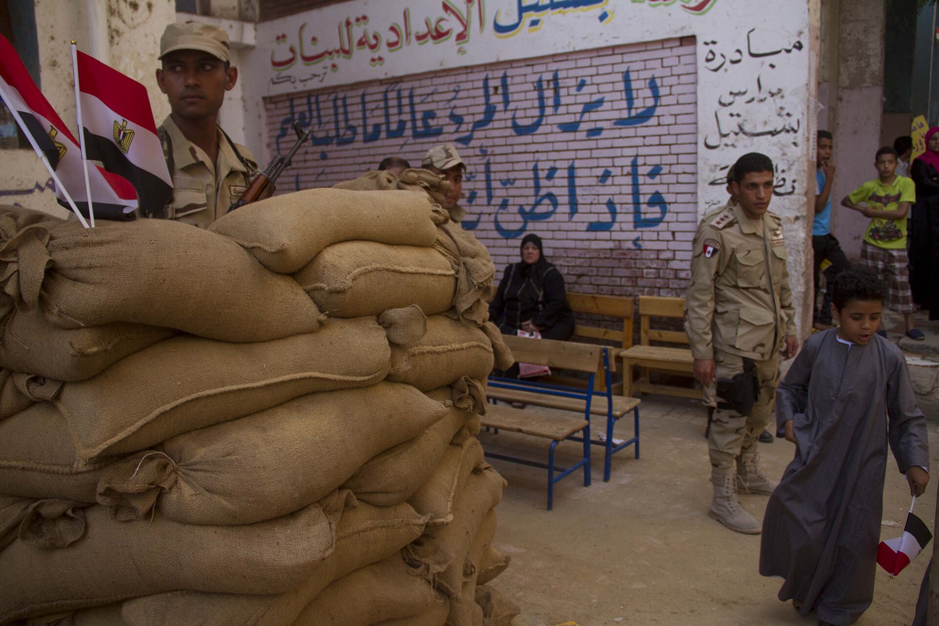 Egyptian soldiers stand beside a sandbag barrier with Egyptian flags outside a school used as a polling station, as a boy walks past holding a small flag