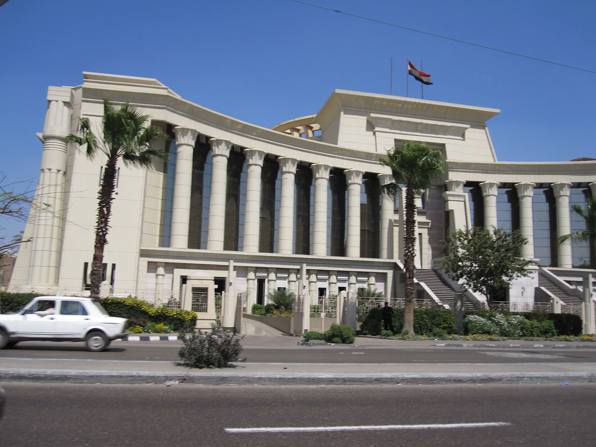 The neo-pharaonic Supreme Constitutional Court building in Cairo with tall lotus-style columns, palm trees, an Egyptian flag and a white car passing
