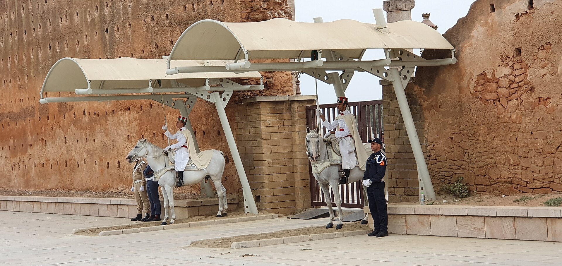 Two mounted royal guards in white uniforms on grey horses under canopies, with a police officer, at the Mausoleum of Mohammed V in Rabat