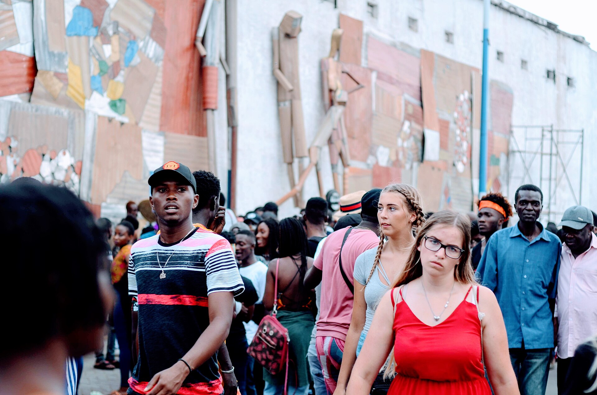 A crowd of young people walks along a street past a wall covered with large collage and sculpture artworks