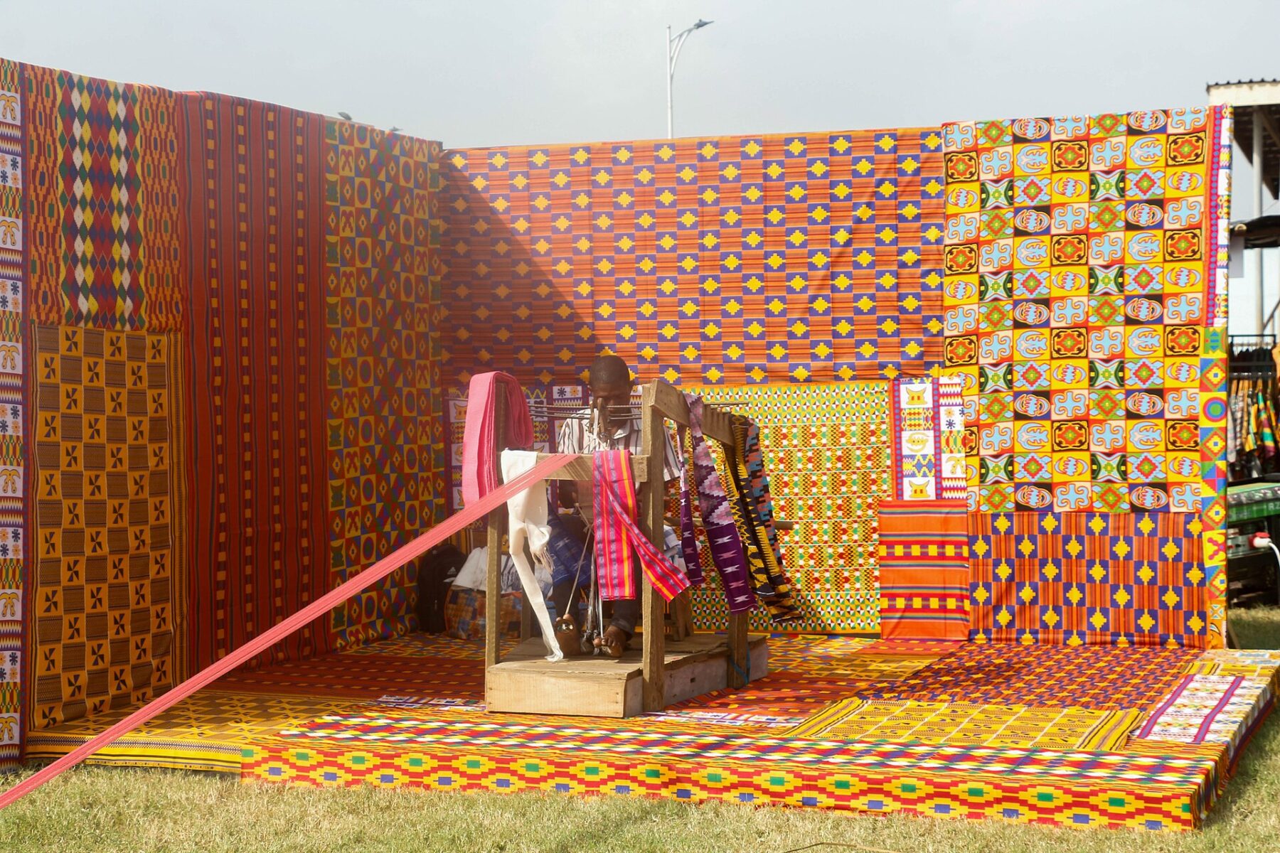 A kente weaver works at a wooden loom inside a booth lined floor to wall with brightly patterned kente cloth in orange, yellow and green