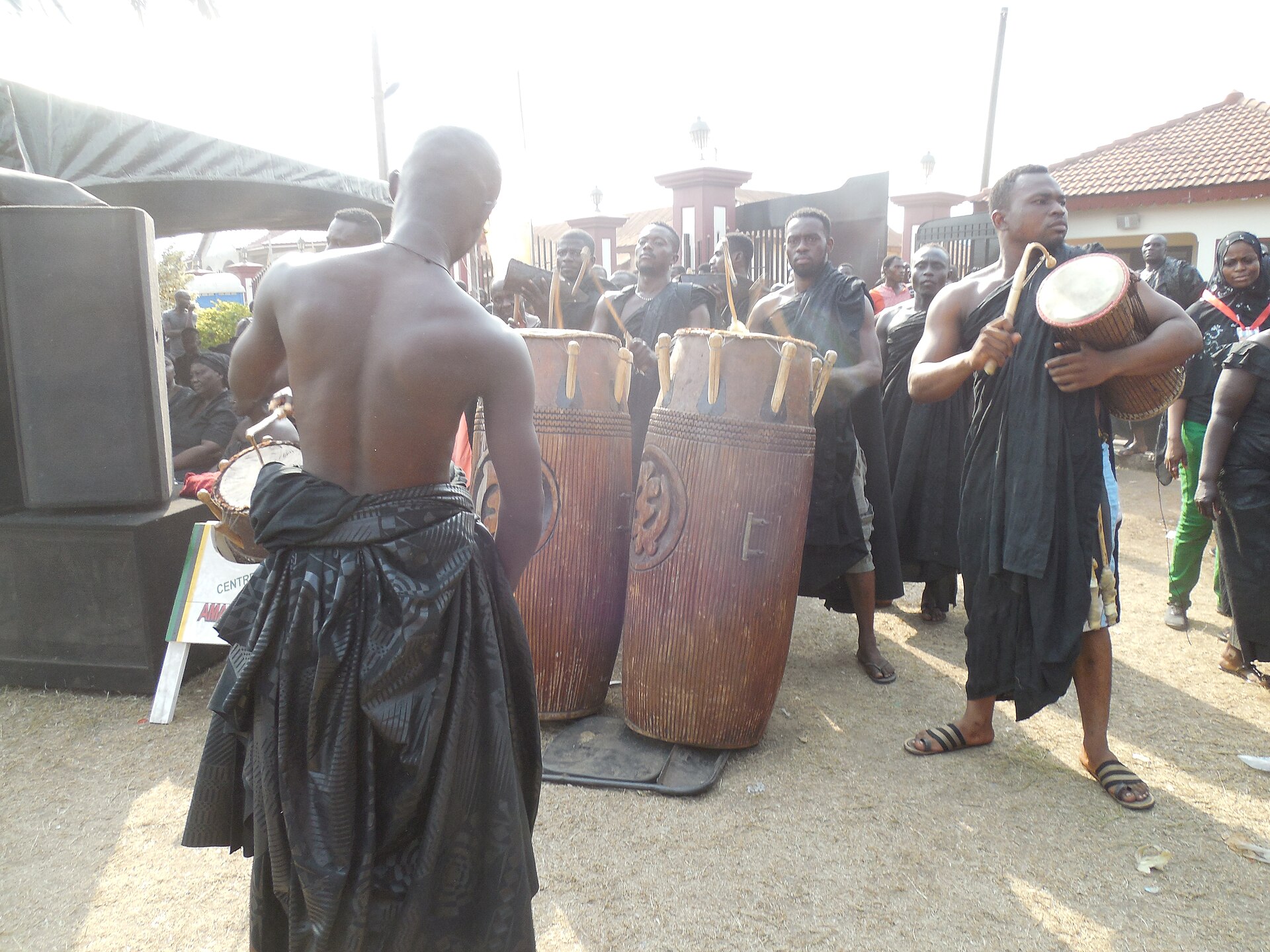 Men in dark cloth wraps play two tall carved drums and hand-held drums outdoors as a crowd looks on