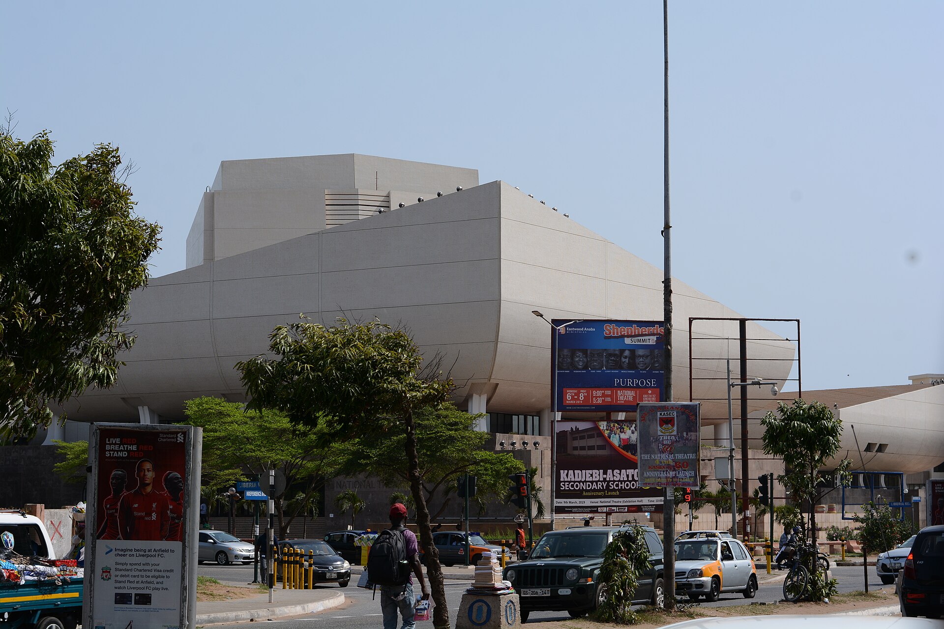 The white, ship-like curved building of the National Theatre of Ghana in Accra behind trees, billboards and street traffic