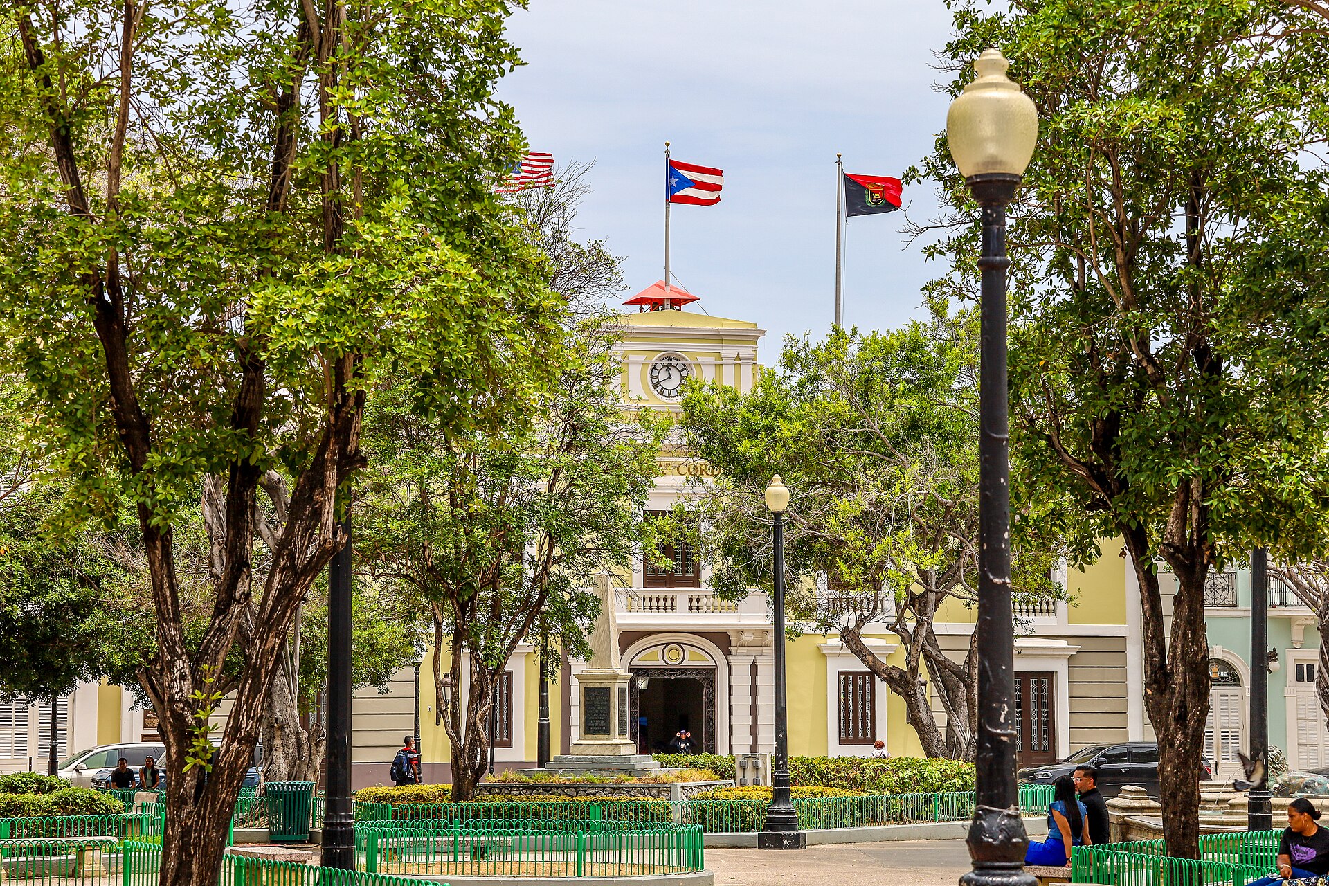 The yellow and cream Ponce City Hall with a clock tower and three flags seen through green trees in Plaza Las Delicias