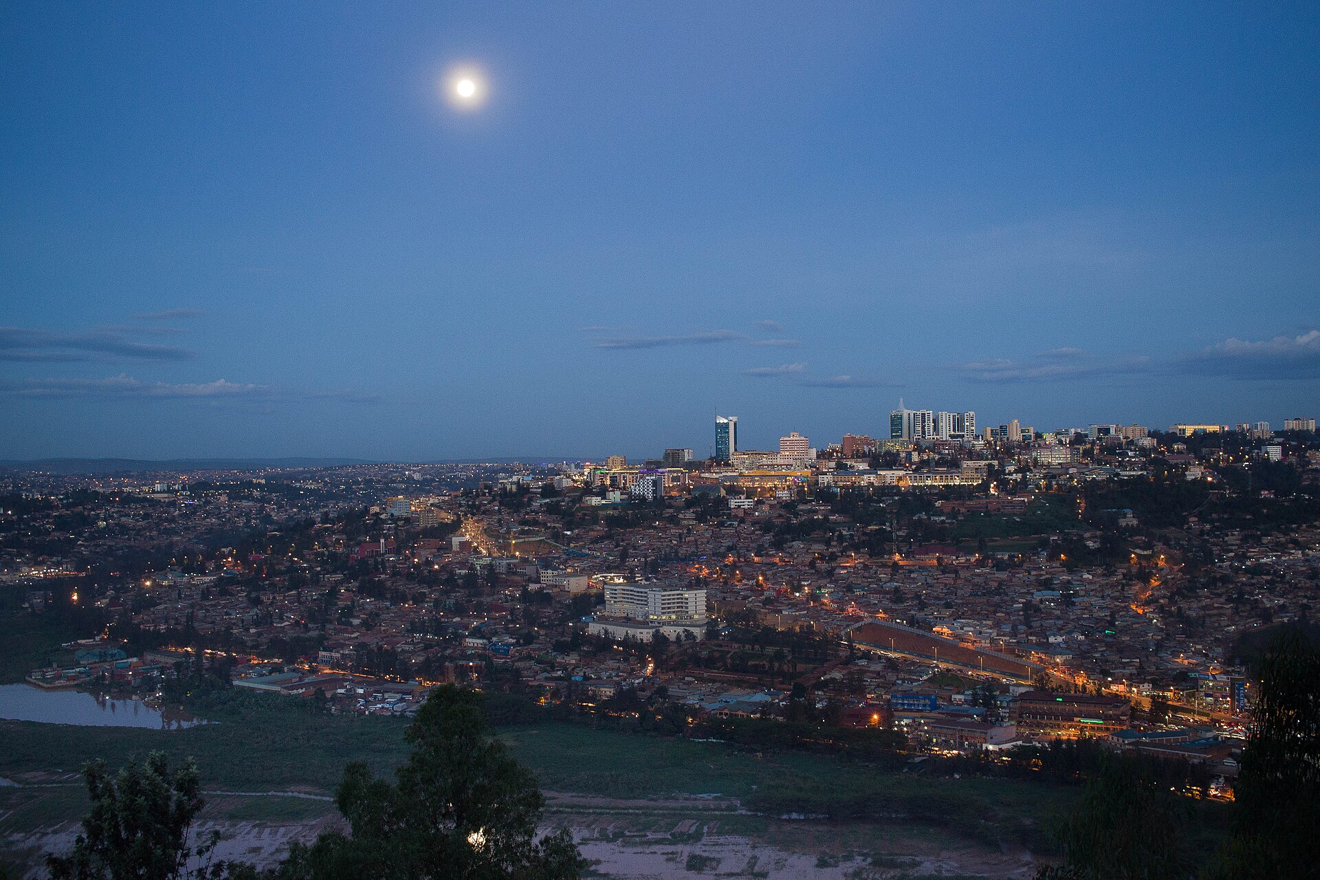 Kigali at dusk seen from a hillside, with city lights spread across the hills, a cluster of office towers and a full moon in a blue sky