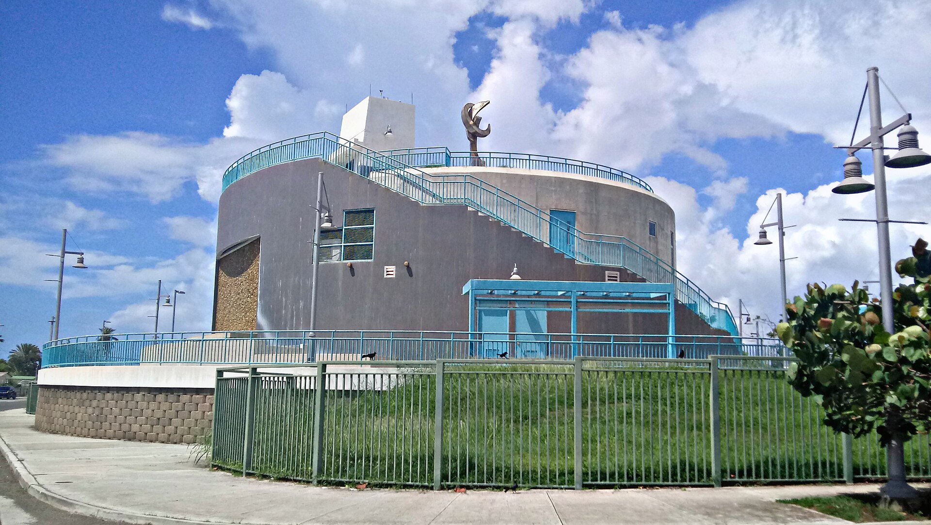 A grey waterfront building with a blue railed staircase and a sculpture on top, seen from a sidewalk under a partly cloudy sky