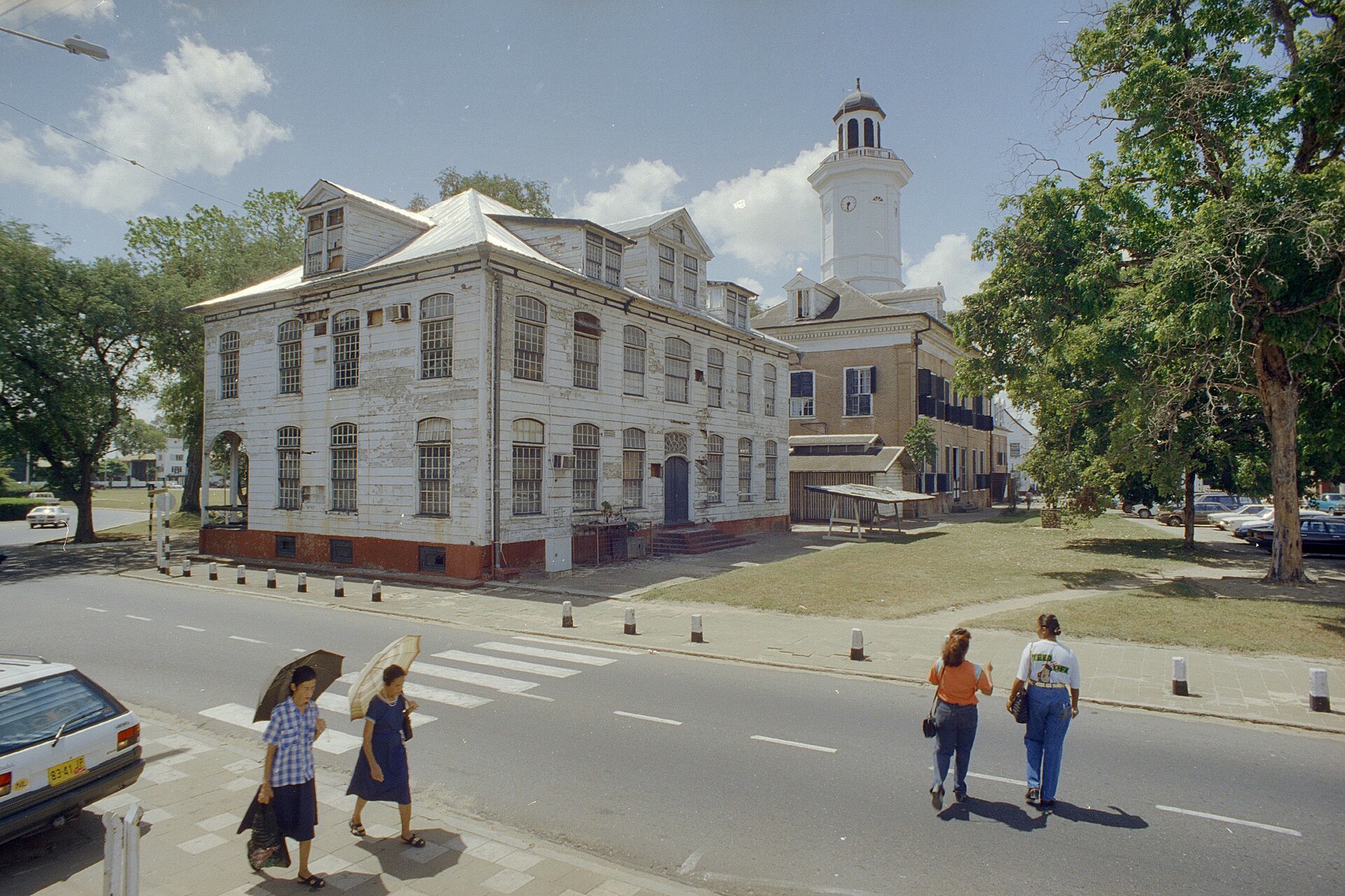 A white wooden colonial building and a white clock tower beside a street in Paramaribo, with pedestrians crossing