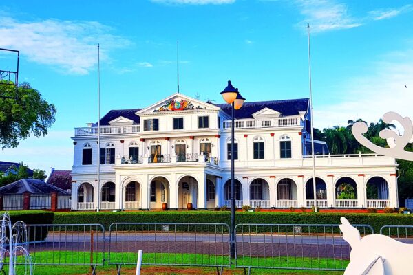 The white colonial Presidential Palace in Paramaribo with dark roof, columns, flagpoles, a lamp post and a lawn under a blue sky