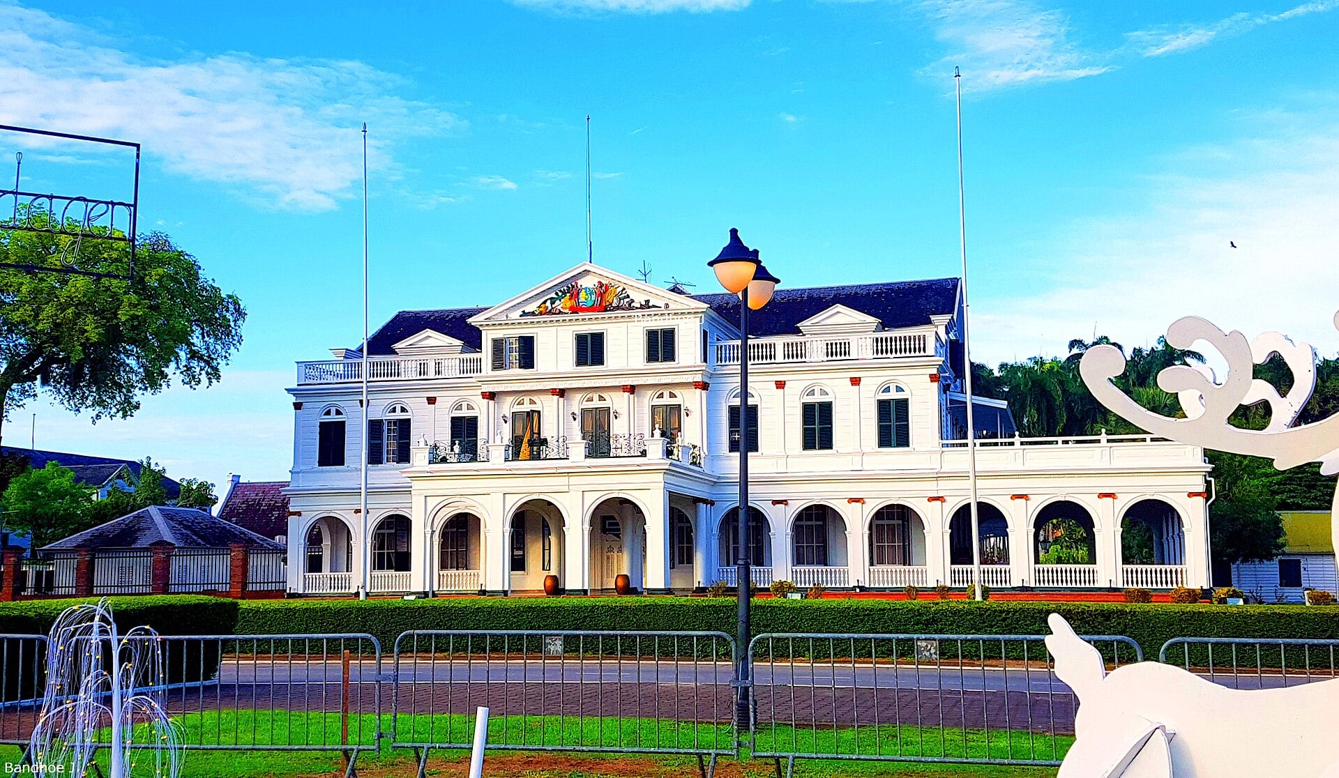 The white colonial Presidential Palace in Paramaribo with dark roof, columns, flagpoles, a lamp post and a lawn under a blue sky