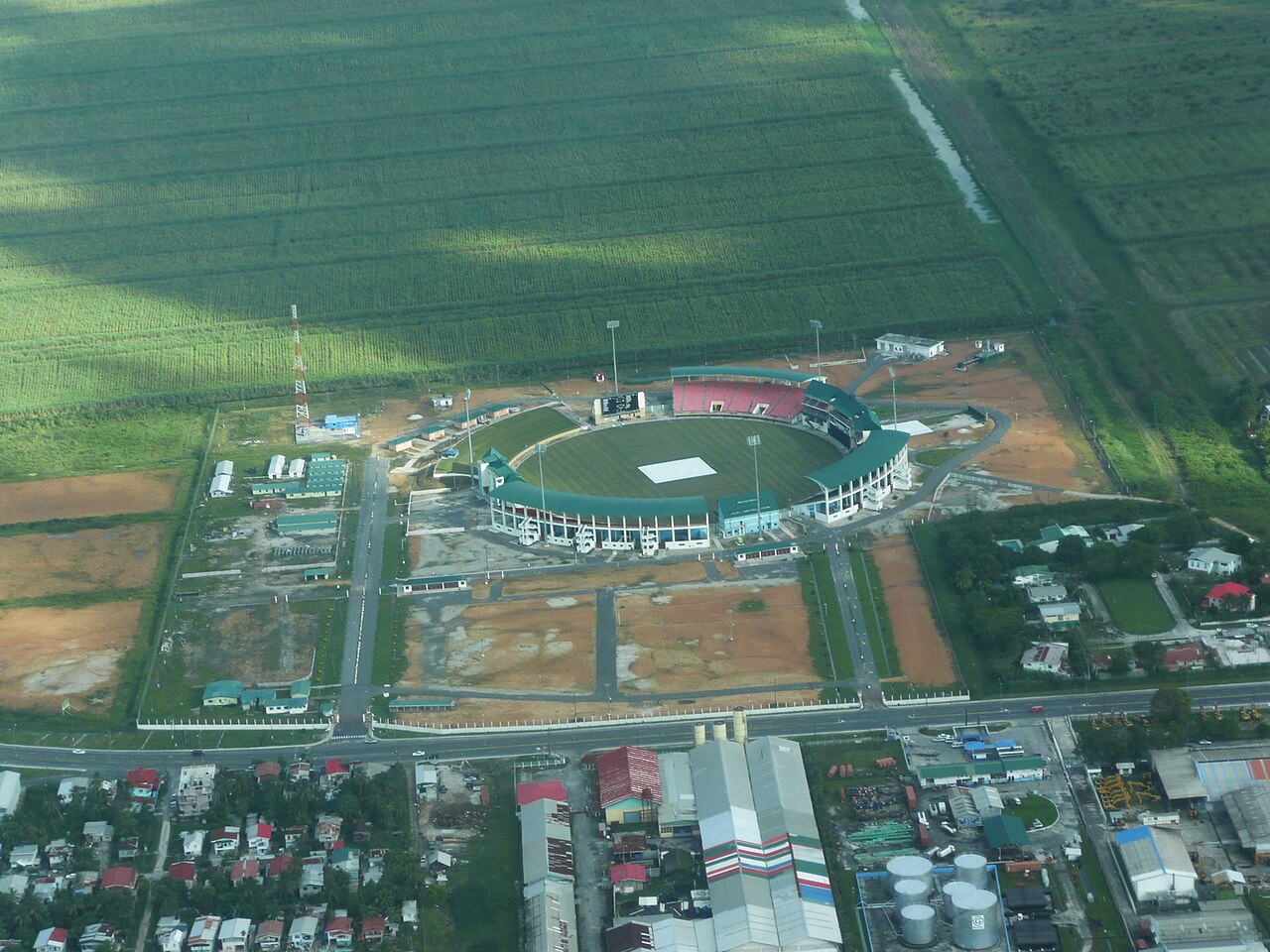 Aerial view of a cricket stadium with green roofs among fields and houses