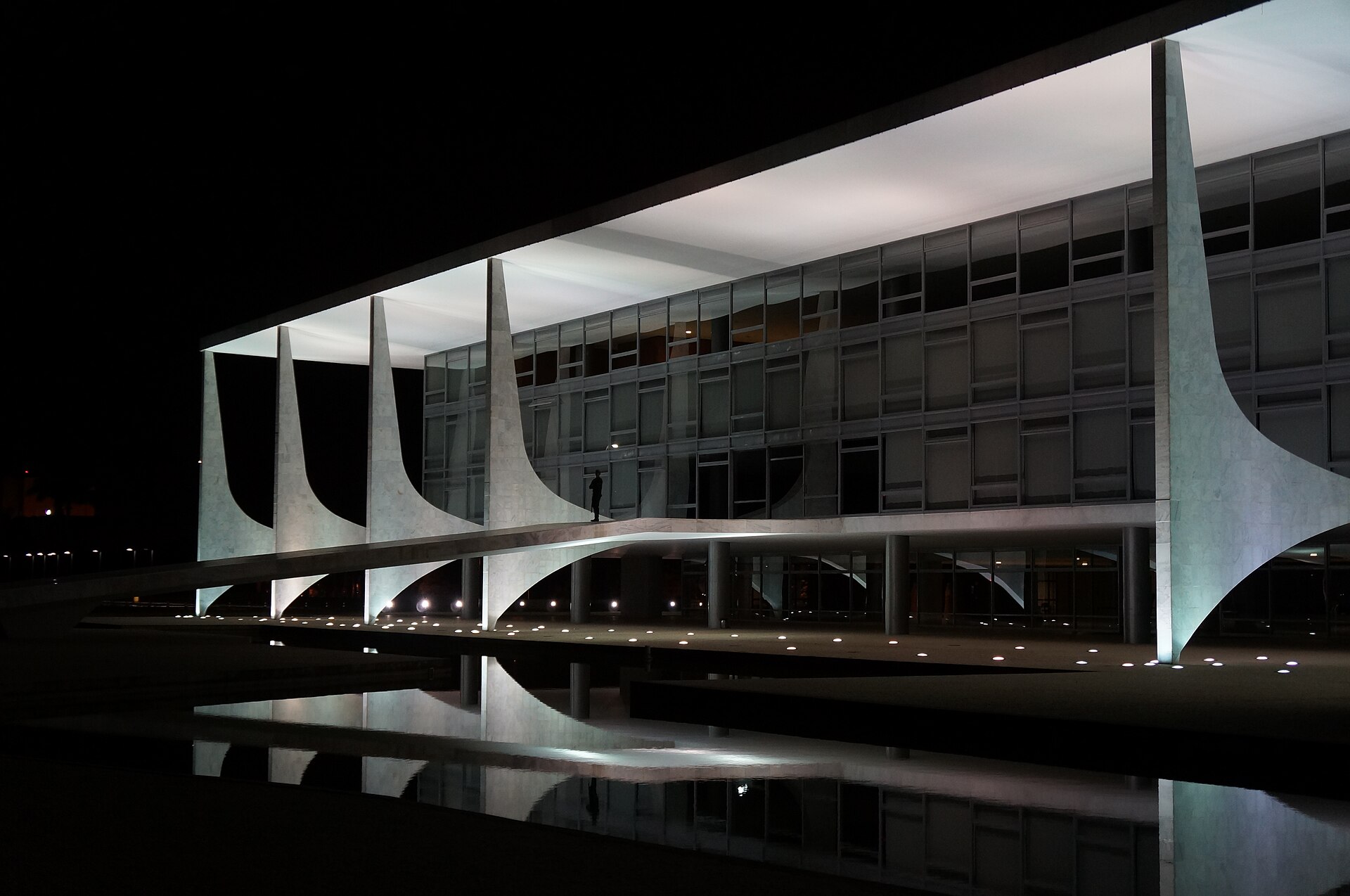 The white columned Planalto Palace in Brasilia lit at night, reflected in a pool, with a lone figure on the ramp