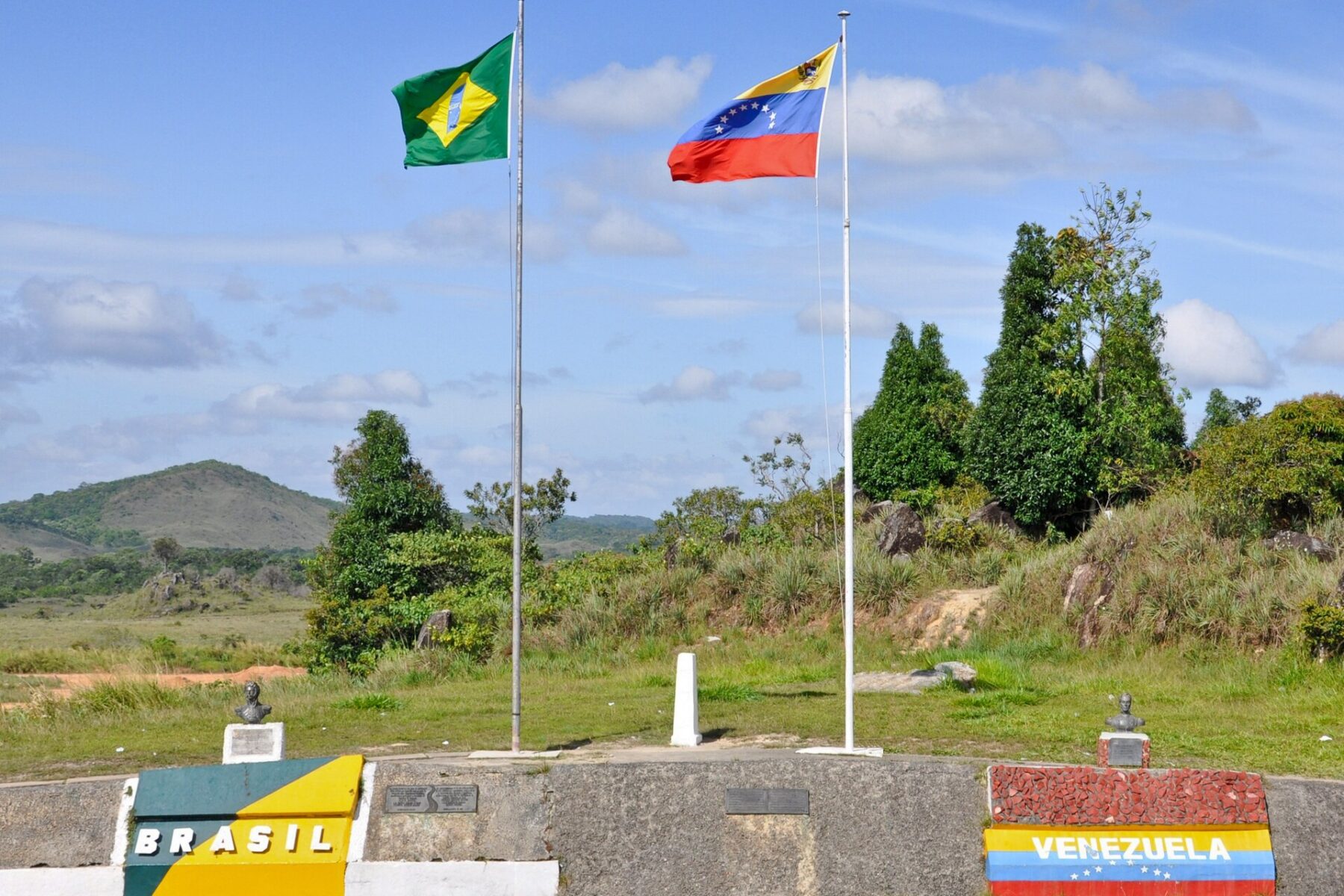 Brazilian and Venezuelan flags on poles at the border marker near Santa Elena de Uairén, with painted national colours on the wall below and green hills behind