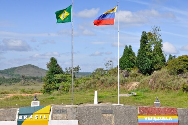 Brazilian and Venezuelan flags on poles at the border marker near Santa Elena de Uairén, with painted national colours on the wall below and green hills behind