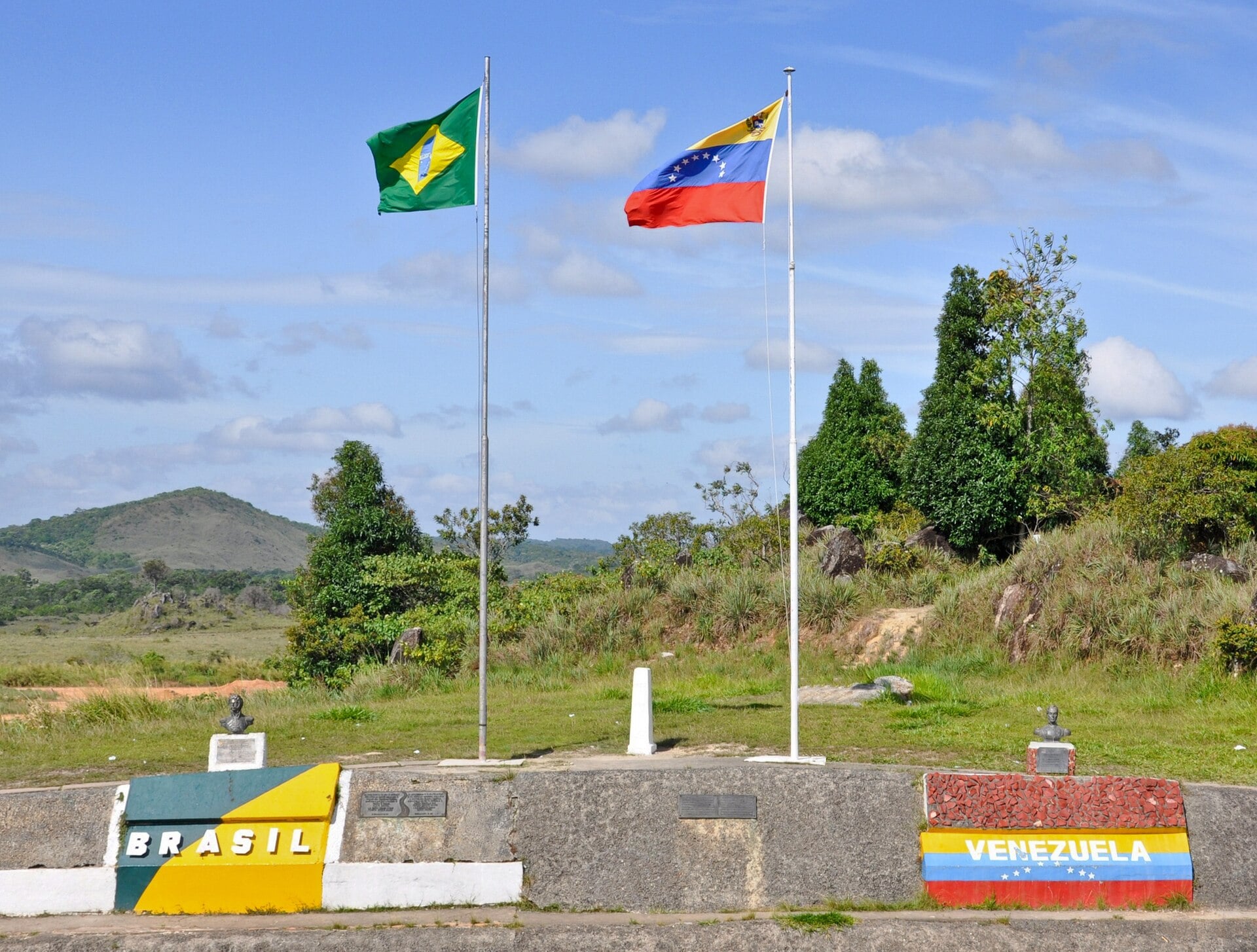 Brazilian and Venezuelan flags on poles at the border marker near Santa Elena de Uairén, with painted national colours on the wall below and green hills behind