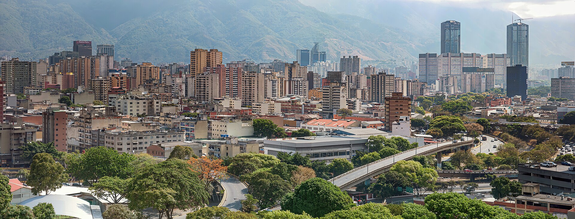 Wide view of Caracas apartment blocks and towers, green trees and a road bridge below the forested Avila mountain