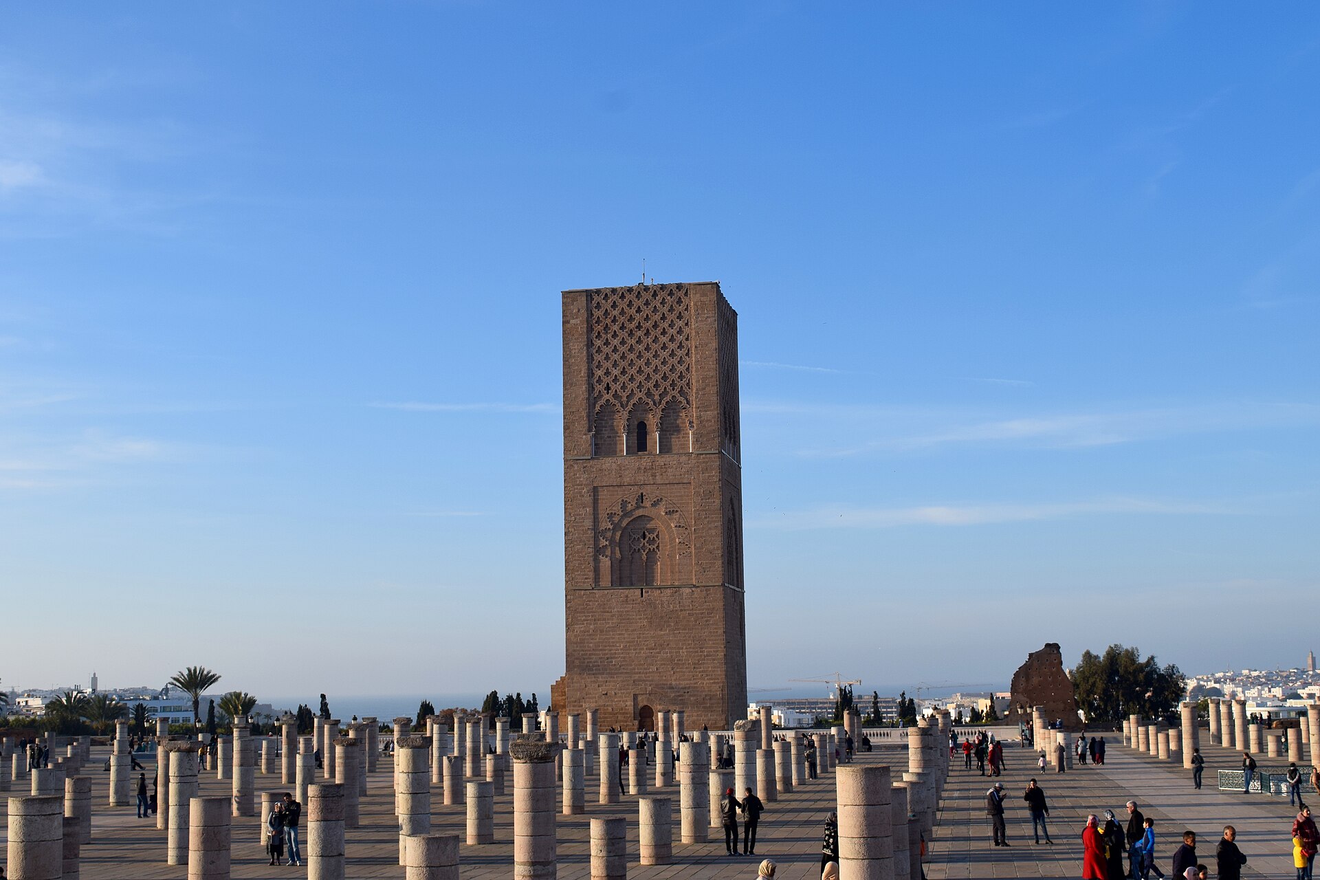 The Hassan Tower in Rabat rising above rows of stone column bases, with visitors walking across the esplanade under a blue sky