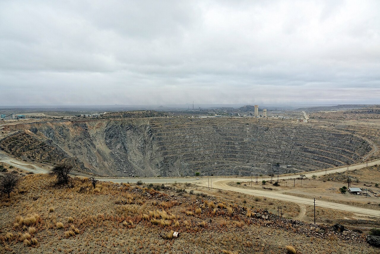 A vast terraced open-pit mine under a grey sky, with a road curving past its edge and industrial buildings on the far rim