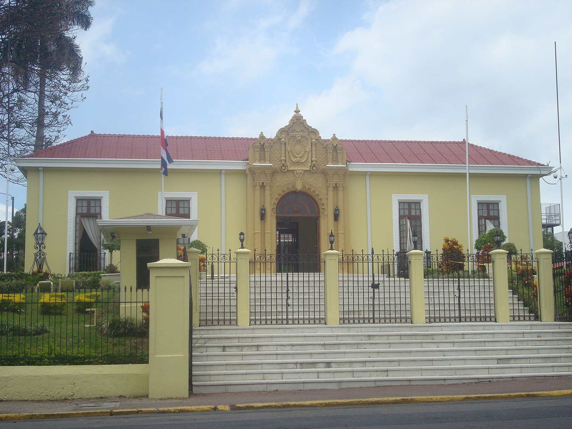 The yellow neo-colonial Casa Amarilla in San José with a red tiled roof, an ornate central doorway, iron fence and steps