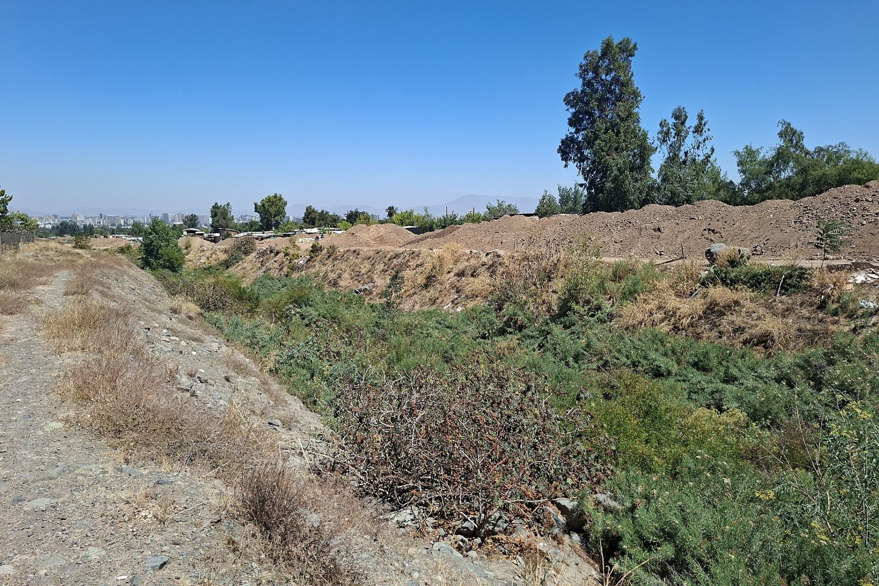 A dry ravine channel with green shrubs and mounds of earth beside it, with Santiago's skyline in the haze in the distance