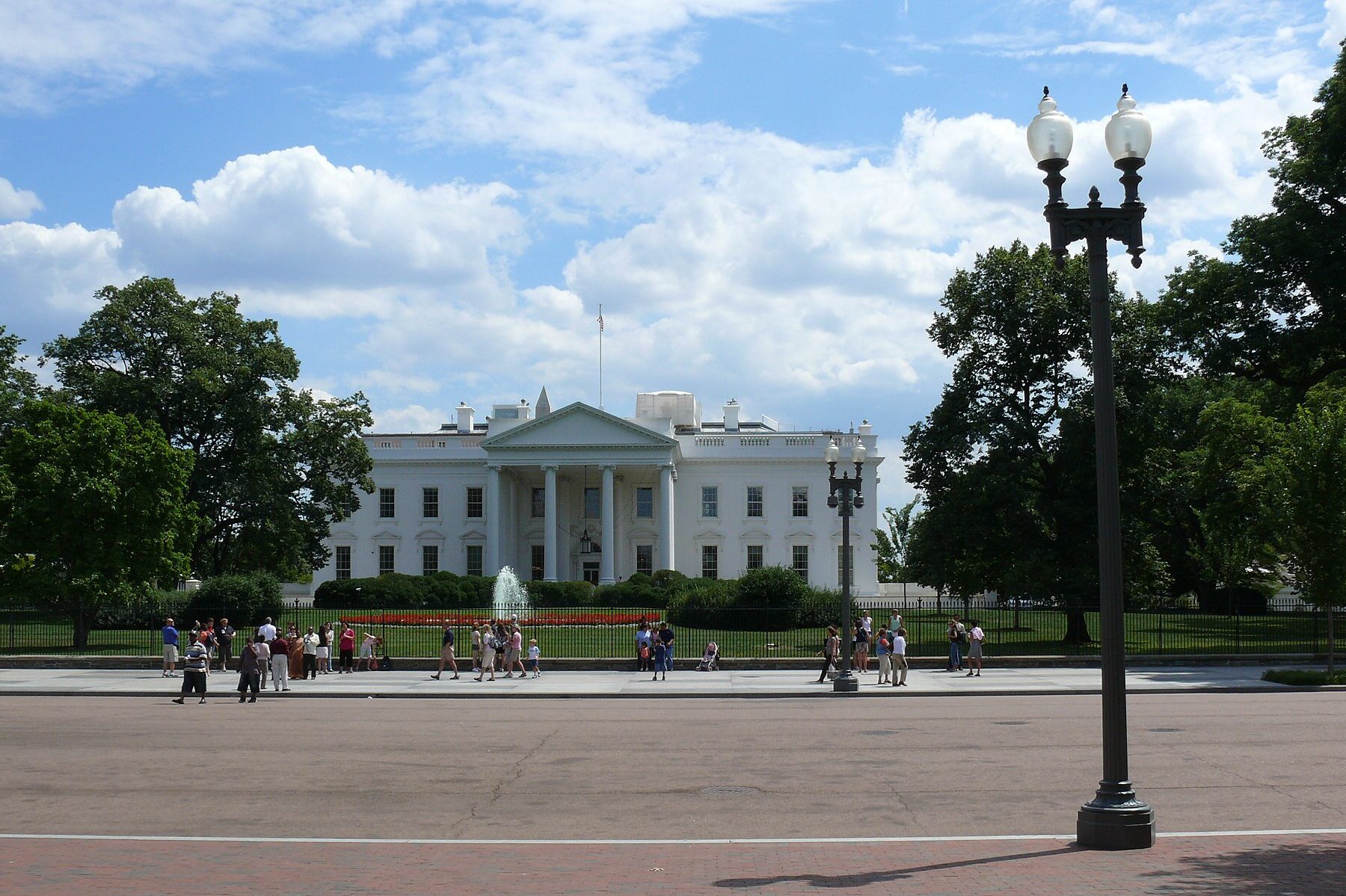 Visitors on the pavement in front of the White House fence on Pennsylvania Avenue under a cloudy sky