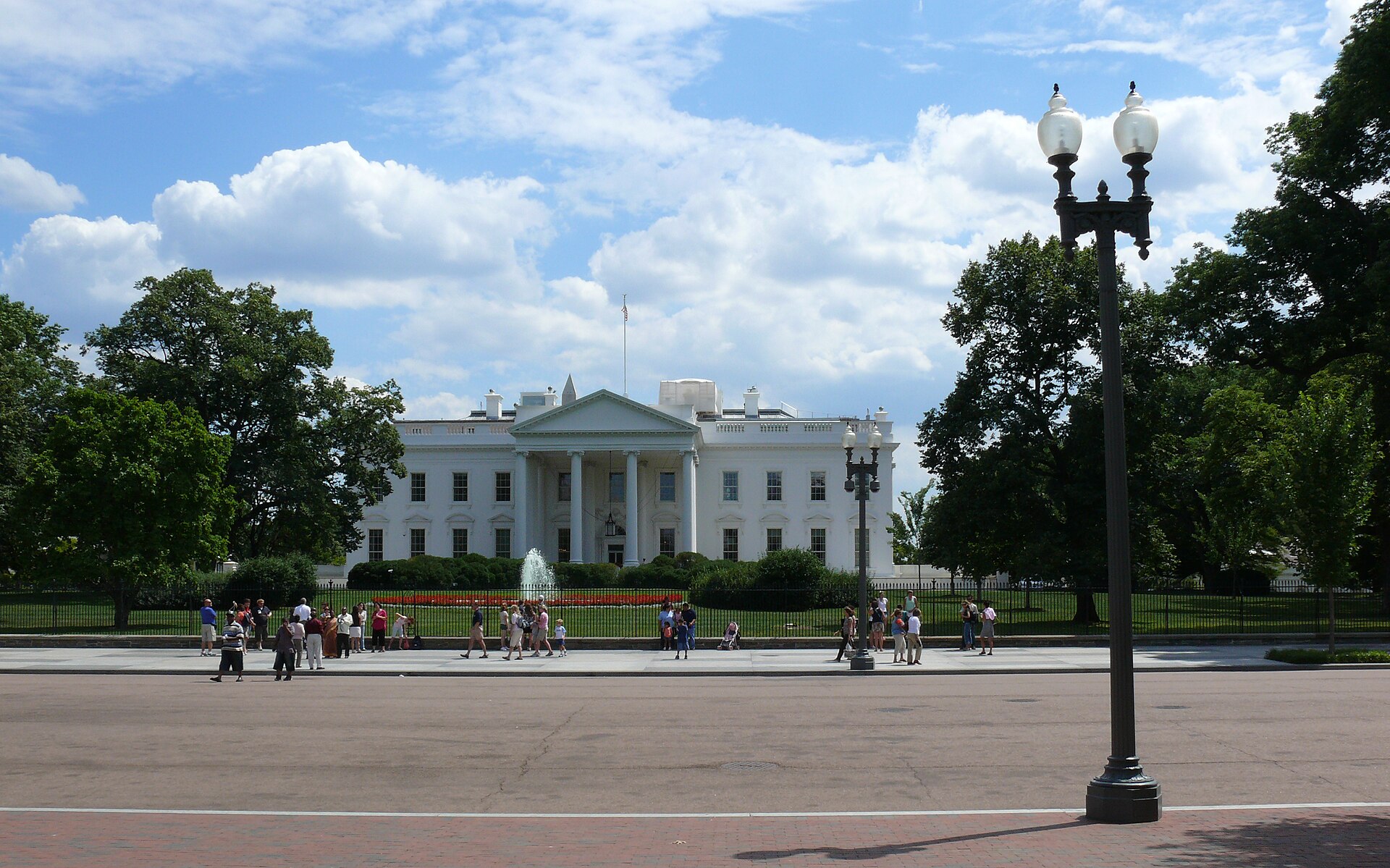 Visitors on the pavement in front of the White House fence on Pennsylvania Avenue under a cloudy sky