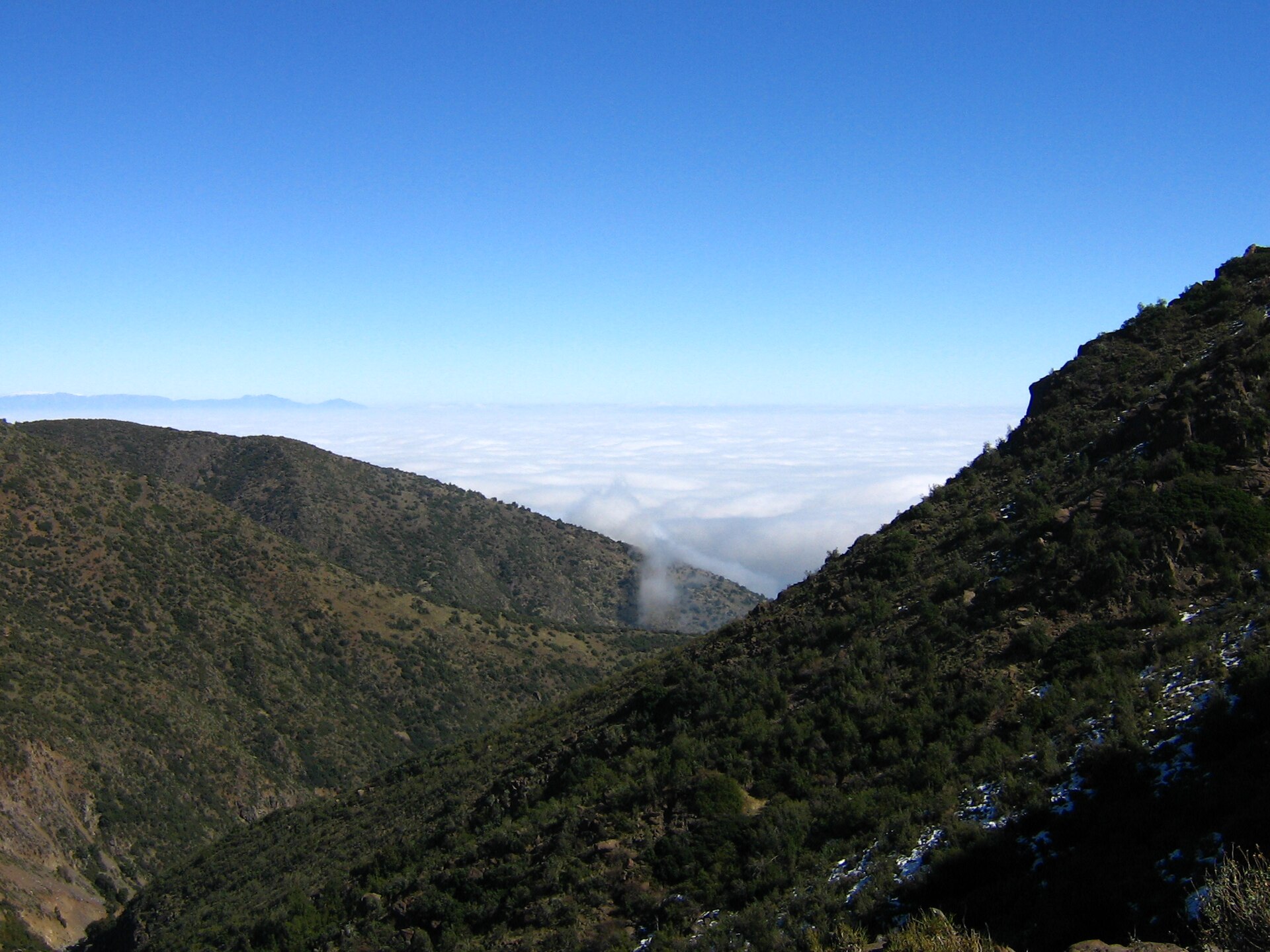 Steep scrub-covered foothill slopes with a layer of cloud filling the valley below and a clear blue sky