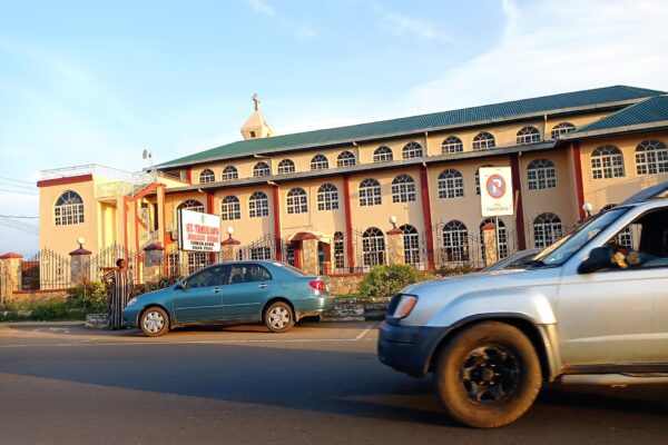 A pink and cream church building with a green roof beside a street in Akure, Ondo State, Nigeria, with cars in the foreground