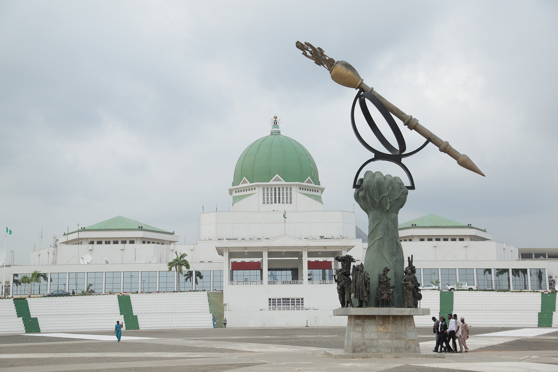 The National Assembly building in Abuja behind a bronze monument of a raised fist holding a ceremonial mace, with a few people walking on the plaza