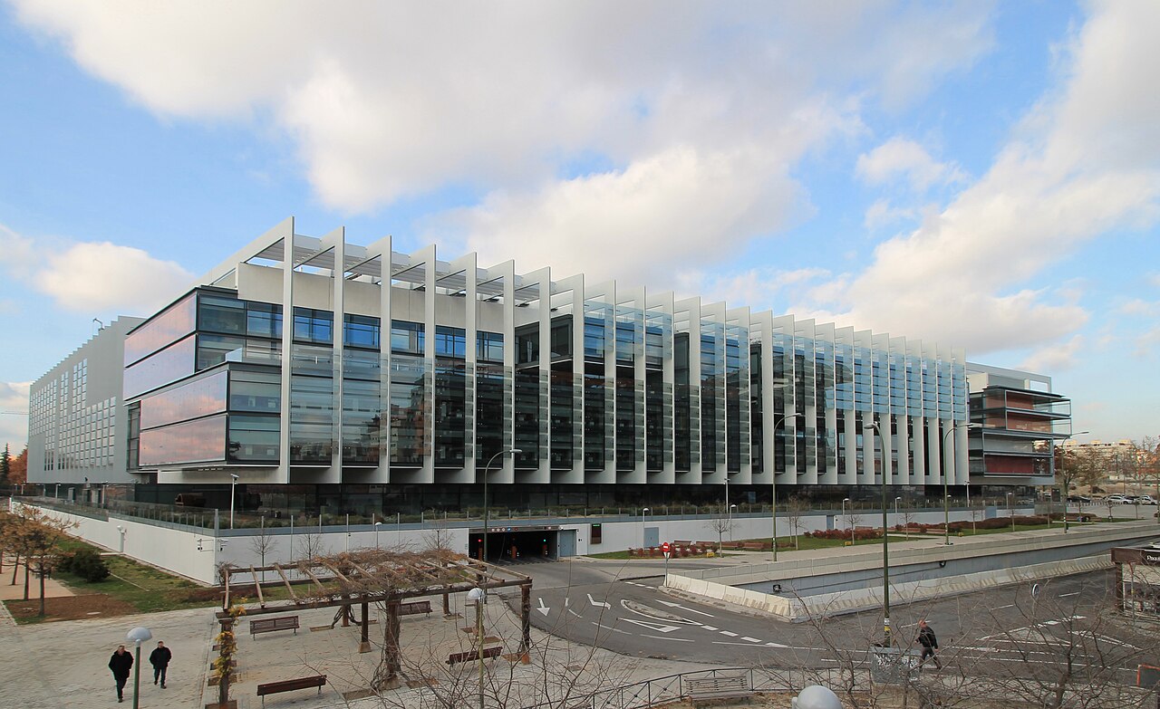 The glass-and-steel Repsol headquarters building in Madrid, with white vertical fins, under a partly cloudy sky